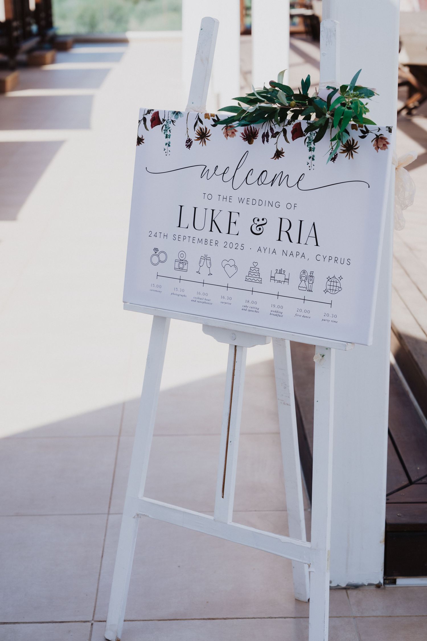 White wedding welcome sign with decorative greenery and timeline details.