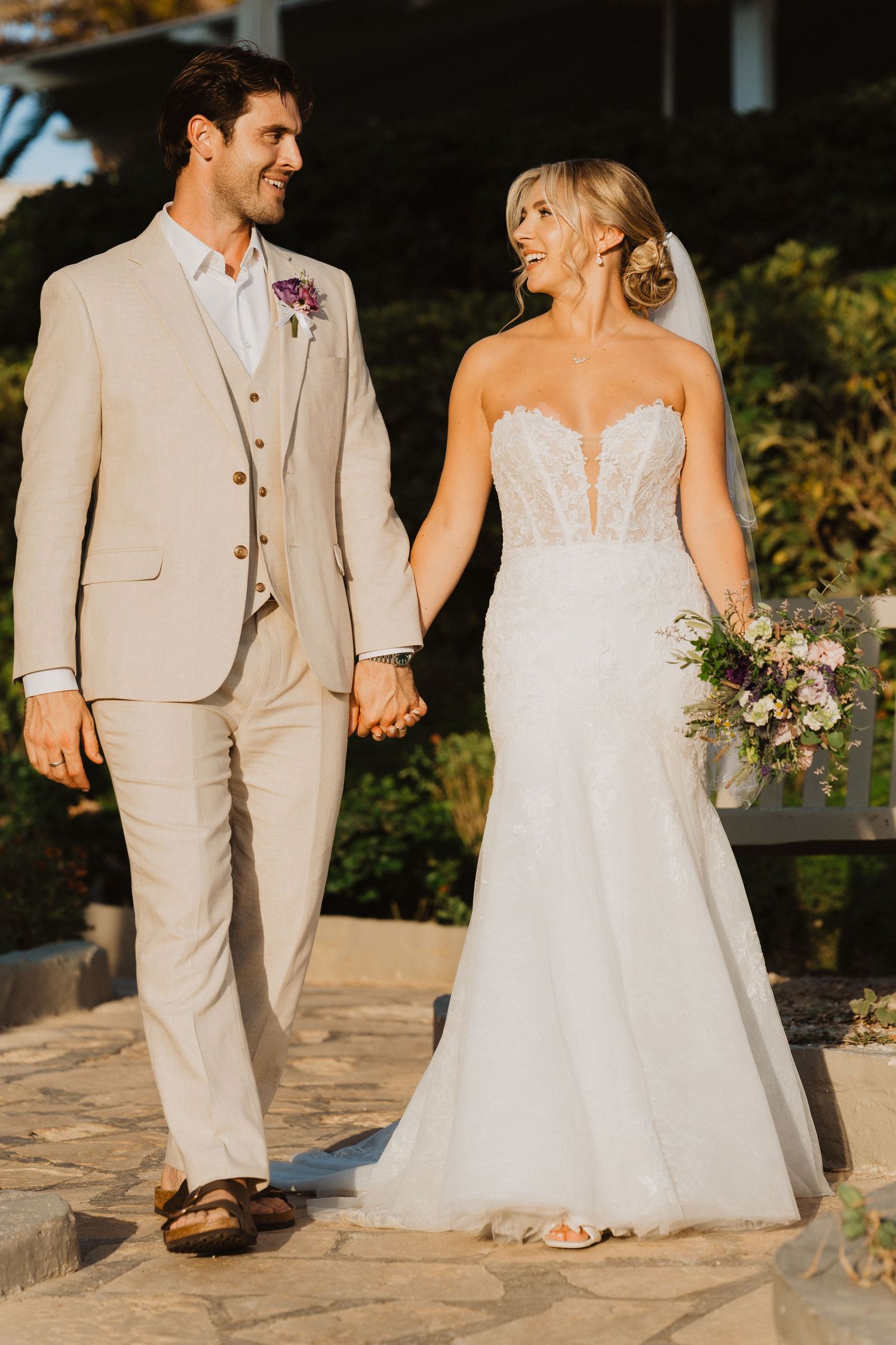 Wedding couple in light beige and white attire walking together on stone pathway.
