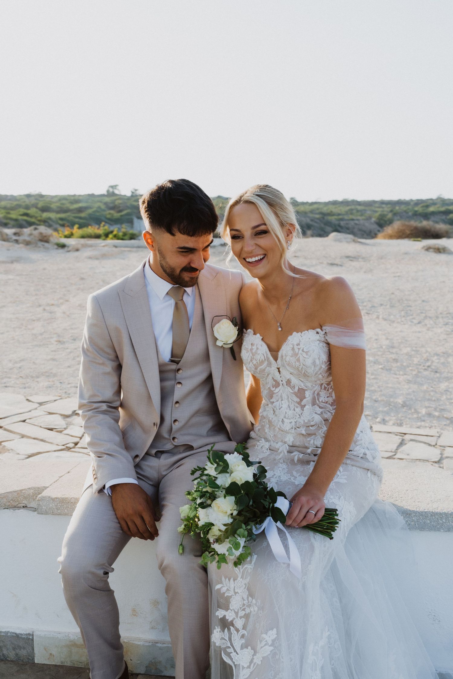 Newlyweds share an intimate moment on a white stone terrace overlooking coastal scenery.