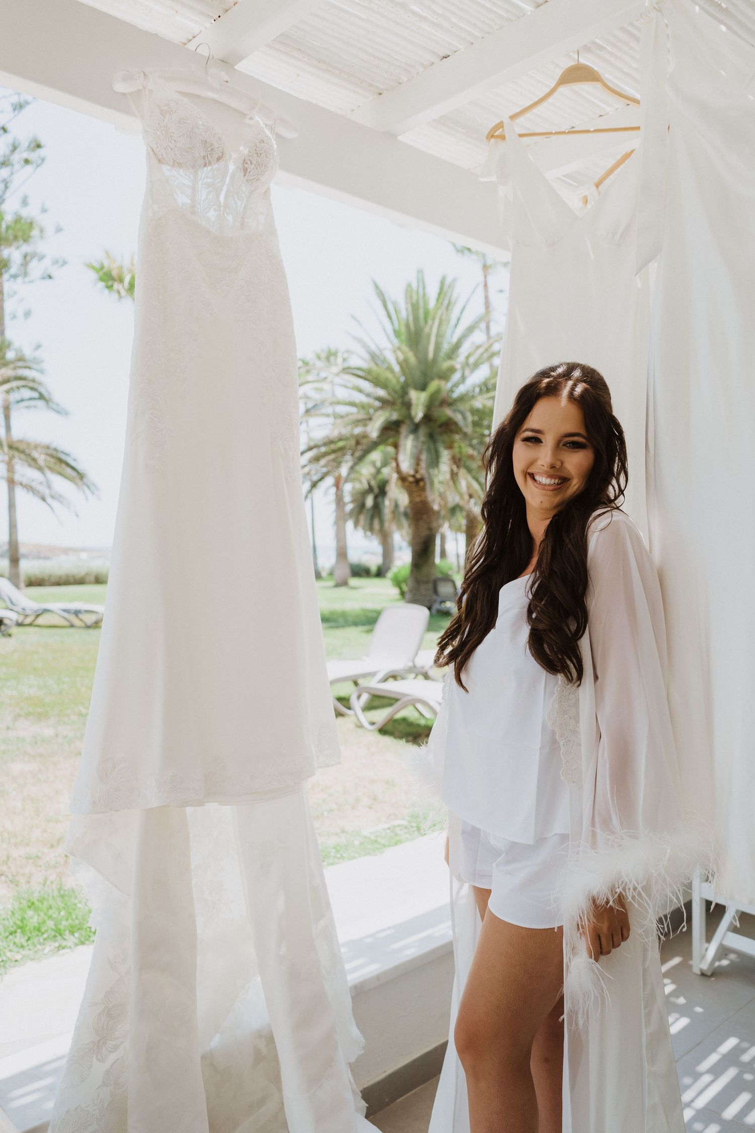 A white wedding dress hangs on display in a bright room with palm trees visible through the window.