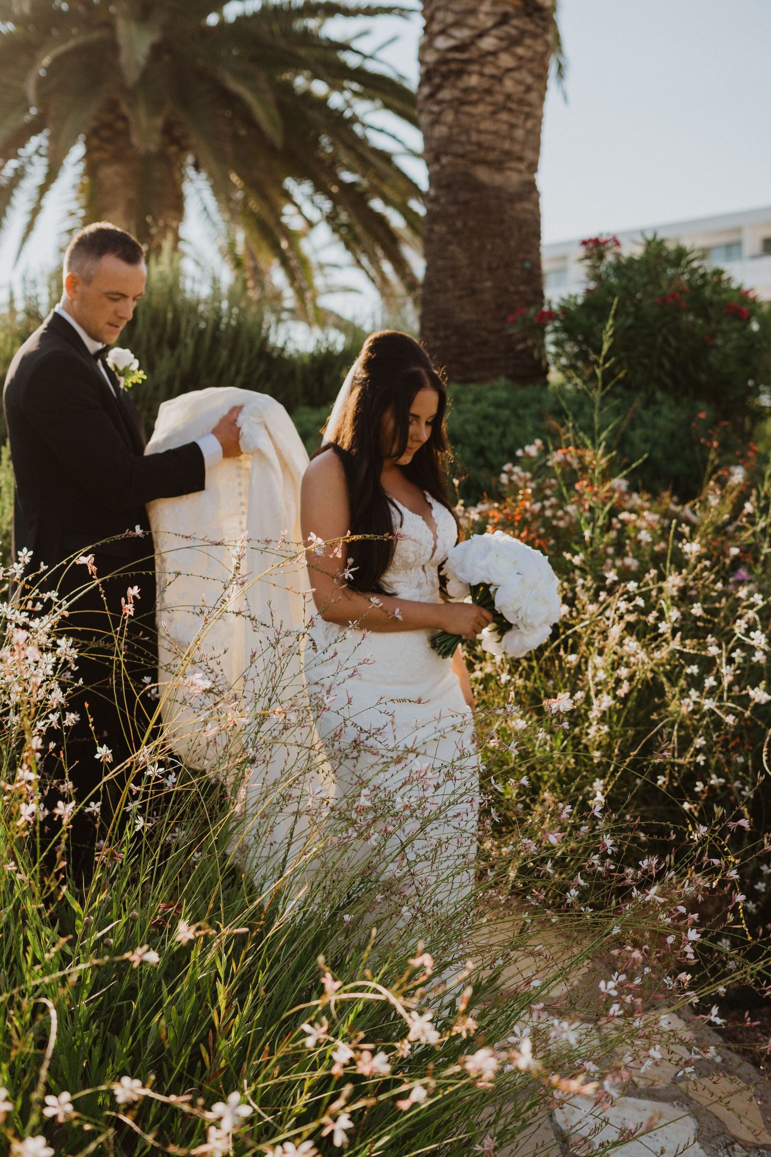 A couple poses in a lush garden setting with palm trees and delicate white flowers during a wedding photoshoot.