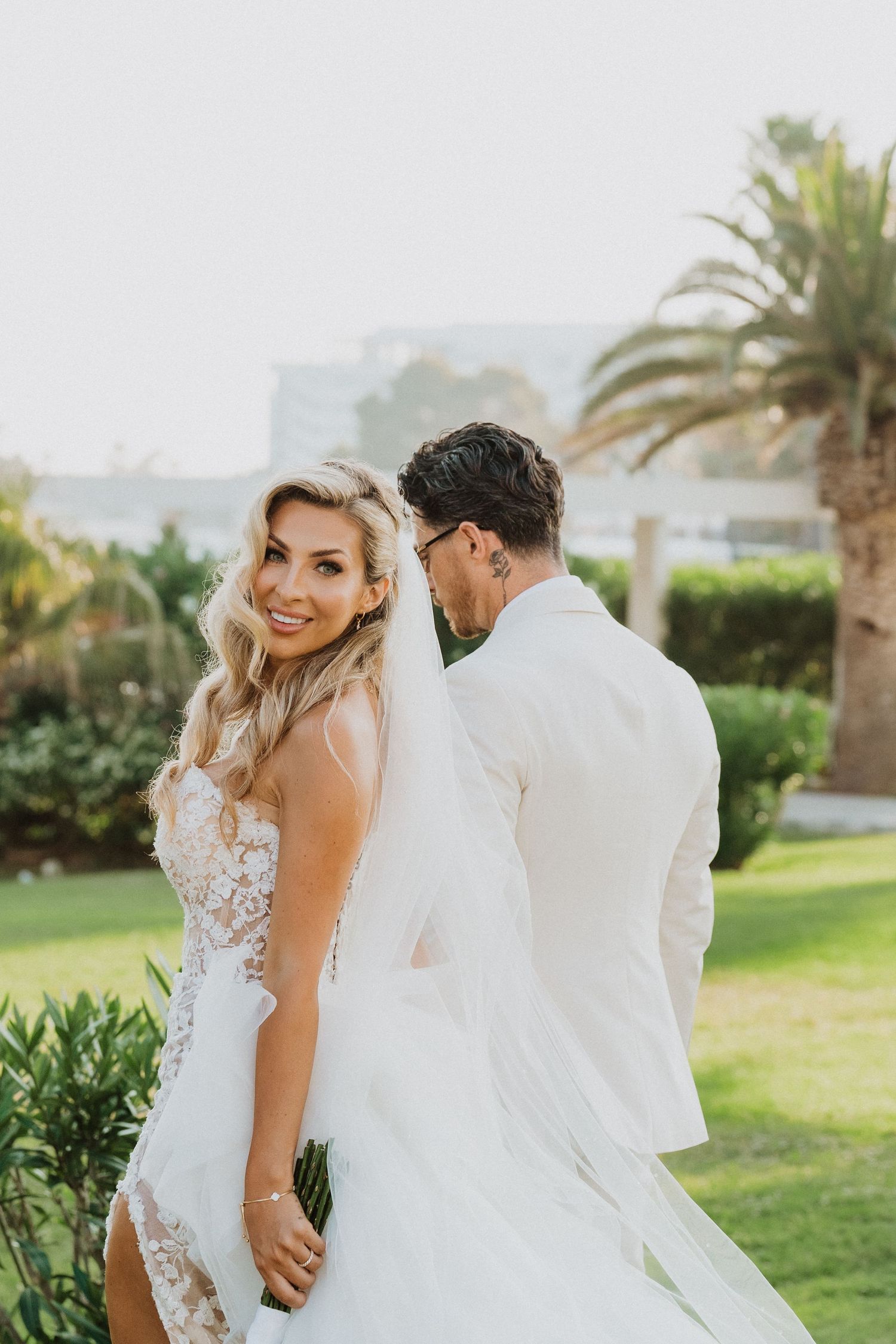 A romantic outdoor wedding scene with a couple in white attire posing in a garden with palm trees.