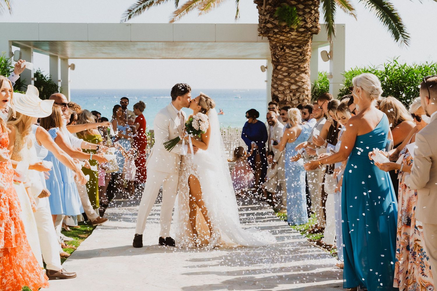 Wedding guests throw colorful confetti at a beach resort celebration as palm trees sway in the background.