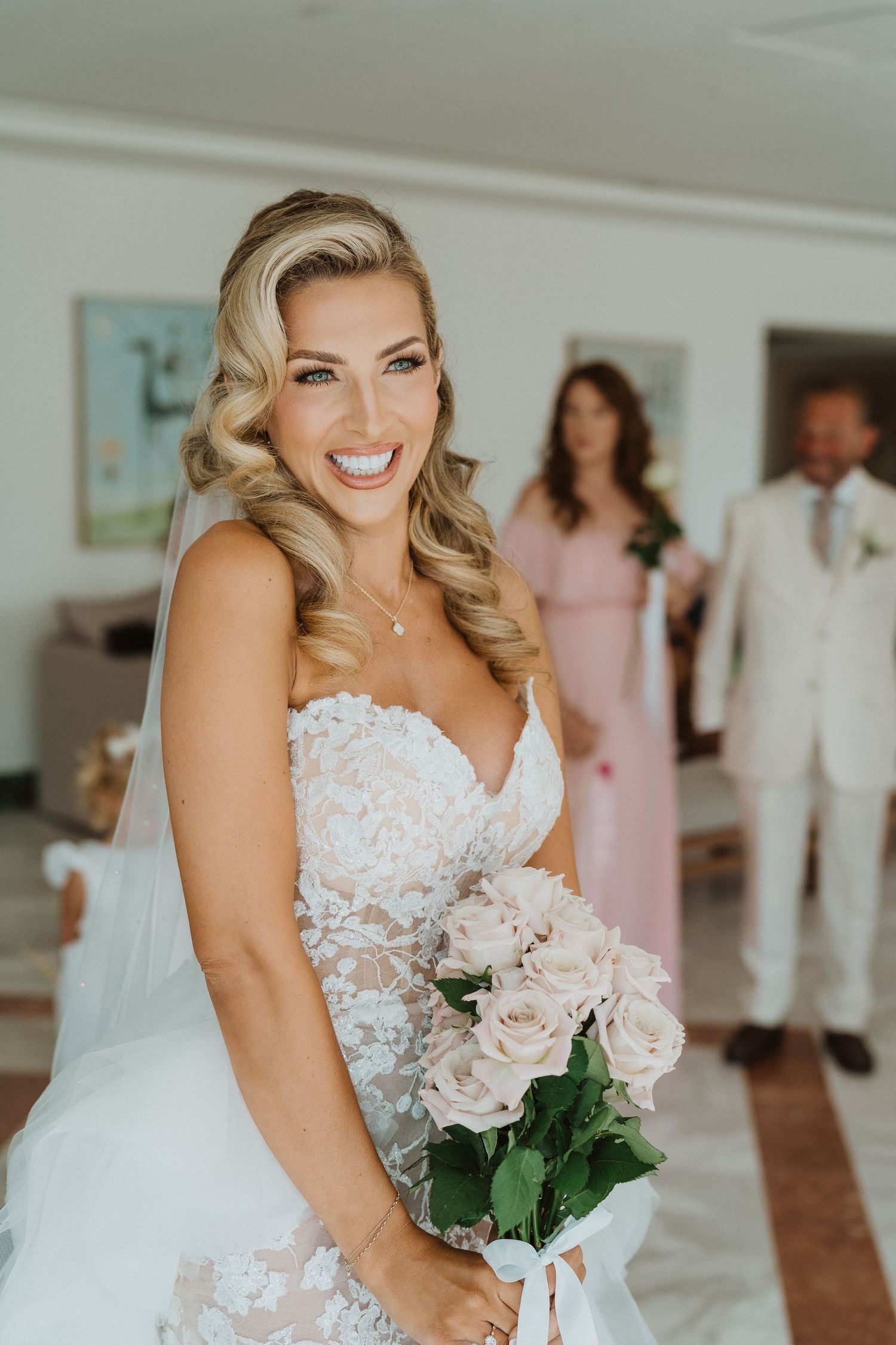 A bride in a white lace wedding dress holds a bouquet of pink roses while guests stand in the background.