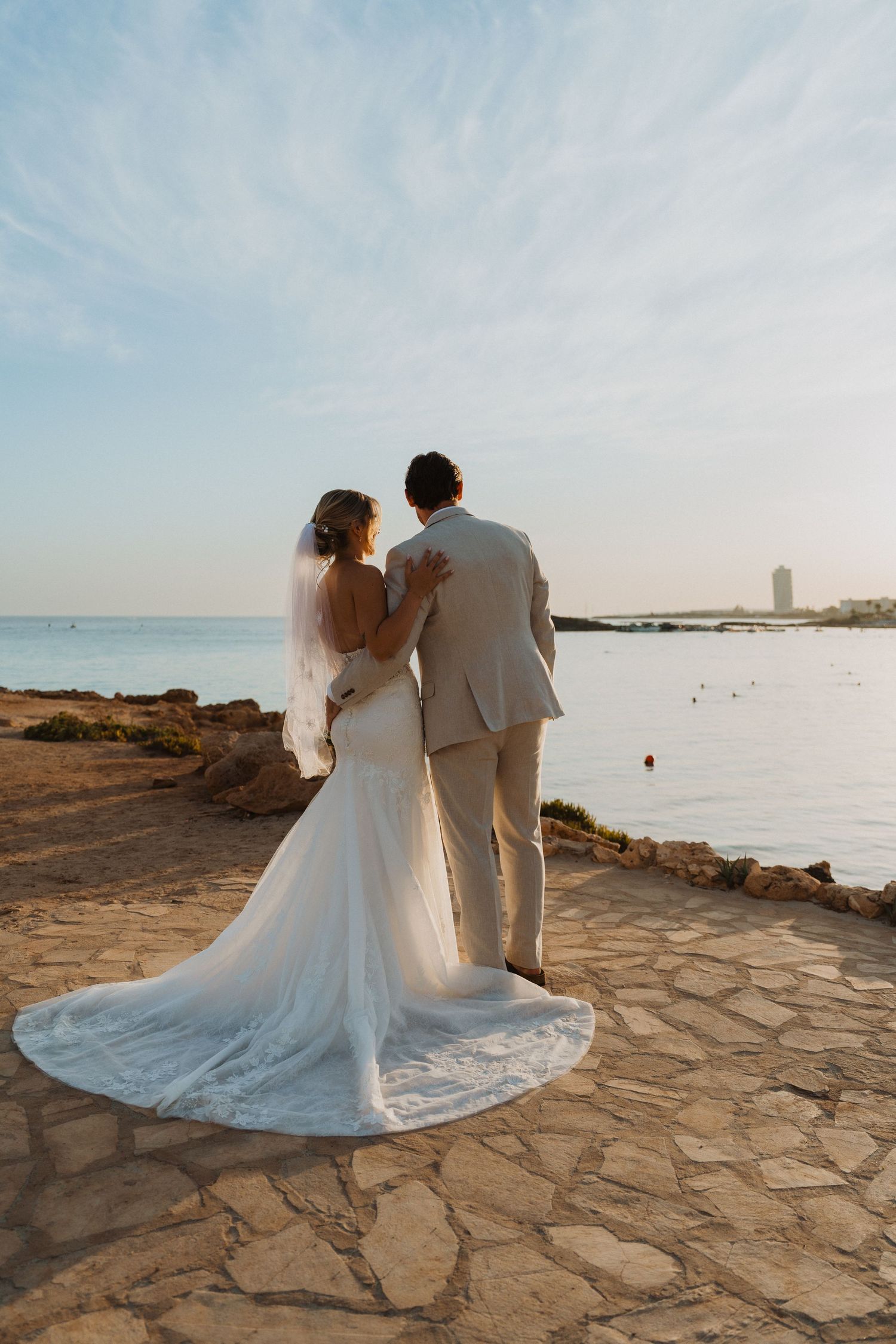 A couple in wedding attire shares an intimate moment on a stone terrace overlooking the ocean at sunset.