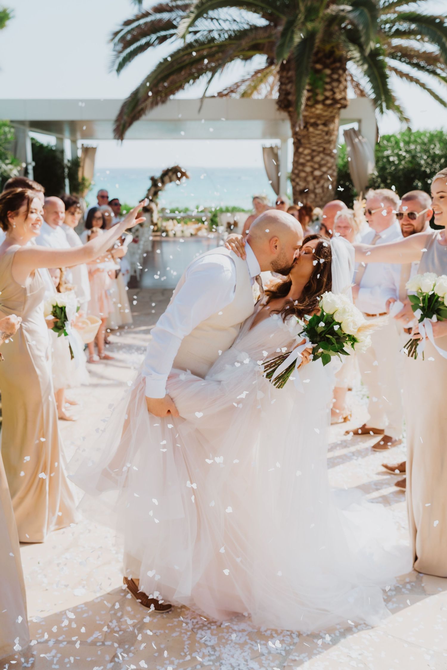 Wedding guests throw white confetti during a joyous celebration under palm trees.