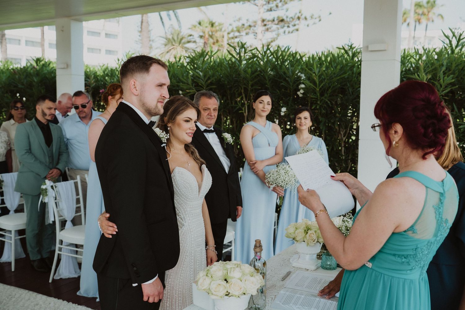 Bride and groom listen to officiant during outdoor wedding ceremony surrounded by guests and bridesmaids.