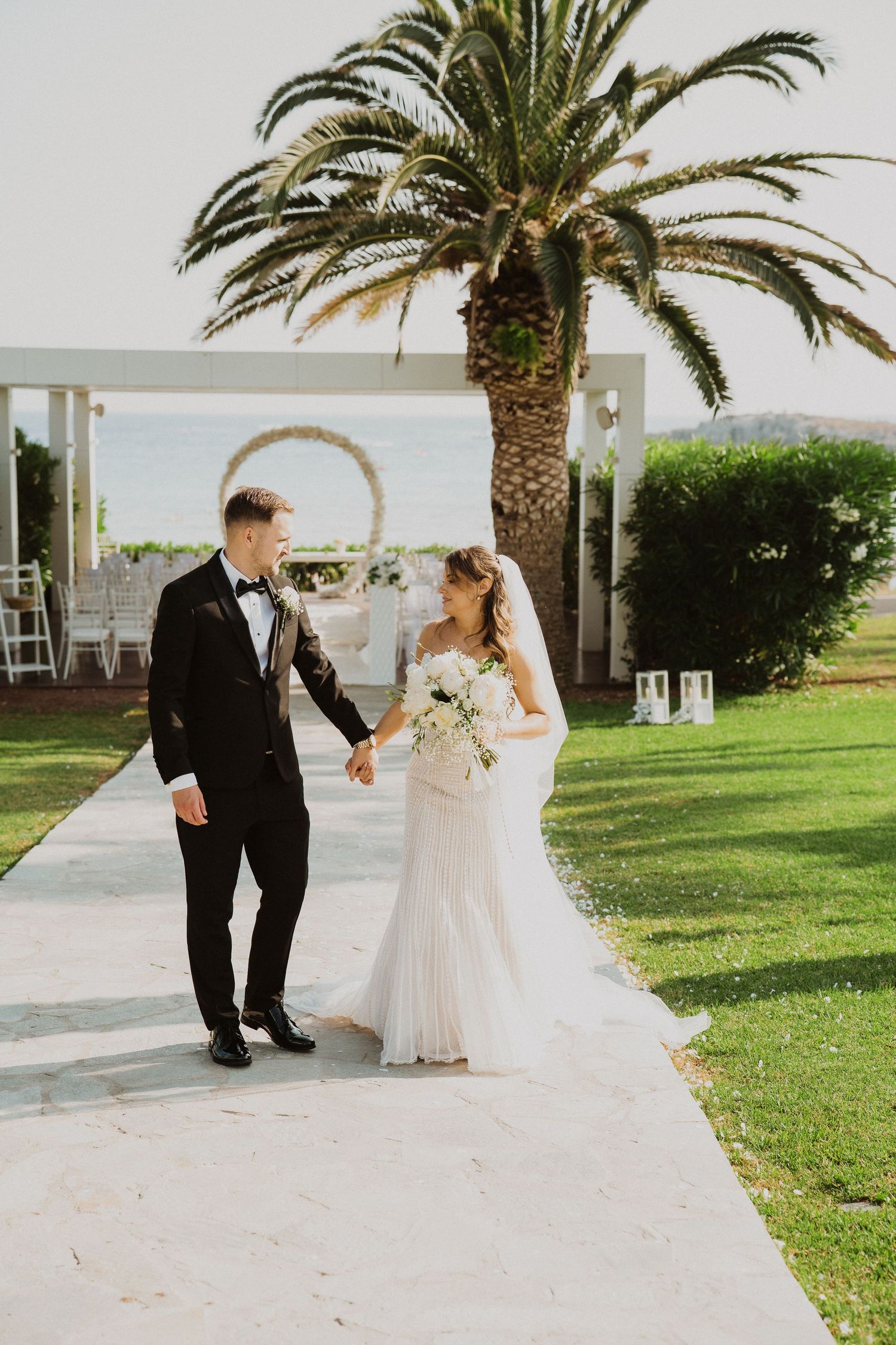 Bride and groom walk hand in hand on a garden path with a palm tree and circular arch backdrop.