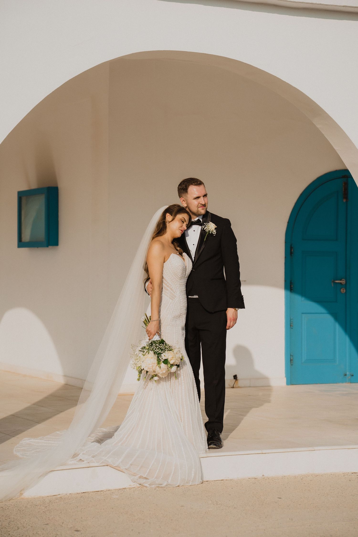 Bride and groom pose under white arch with turquoise door, bride in lace gown with veil, groom in black tuxedo.