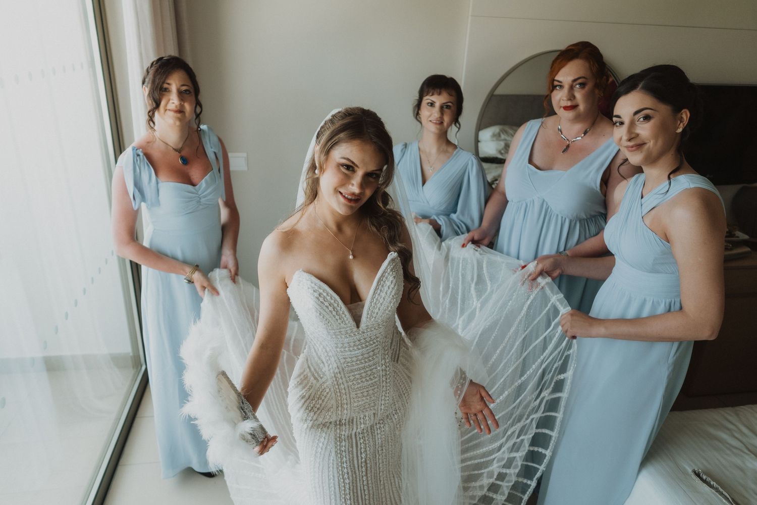 Bride in beaded gown surrounded by four bridesmaids in light blue dresses near a window.