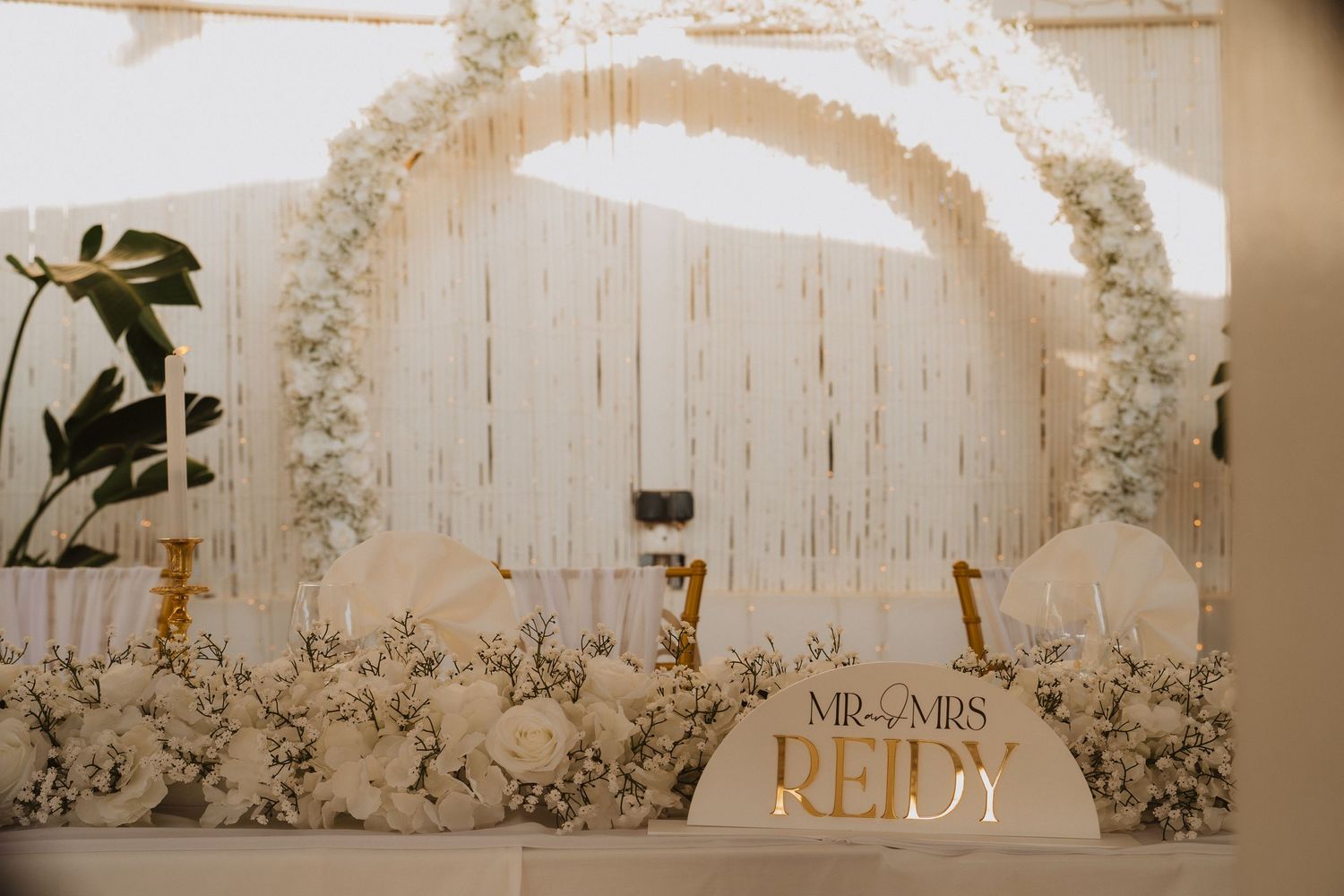 Elegant wedding reception backdrop reading Mr and Mrs Reidy with white floral arch and gold candle holders.
