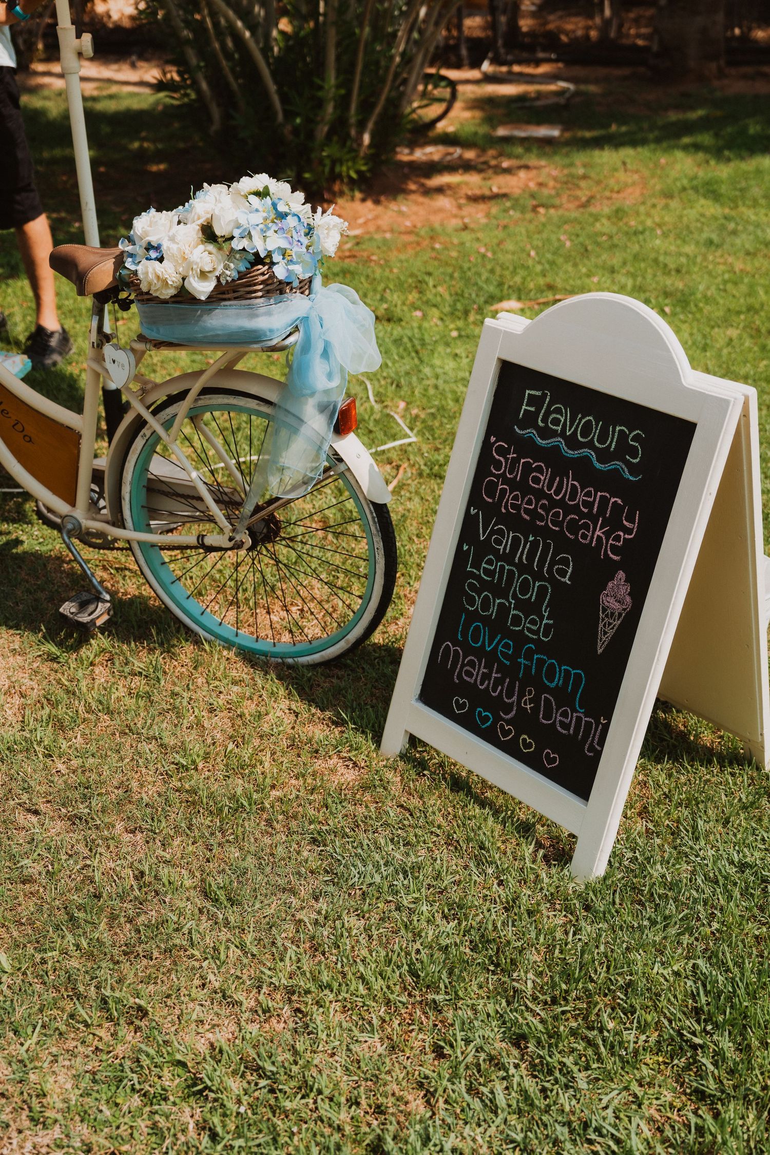 Vintage teal bicycle with floral basket beside a chalkboard sign listing ice cream flavors at an outdoor wedding reception.