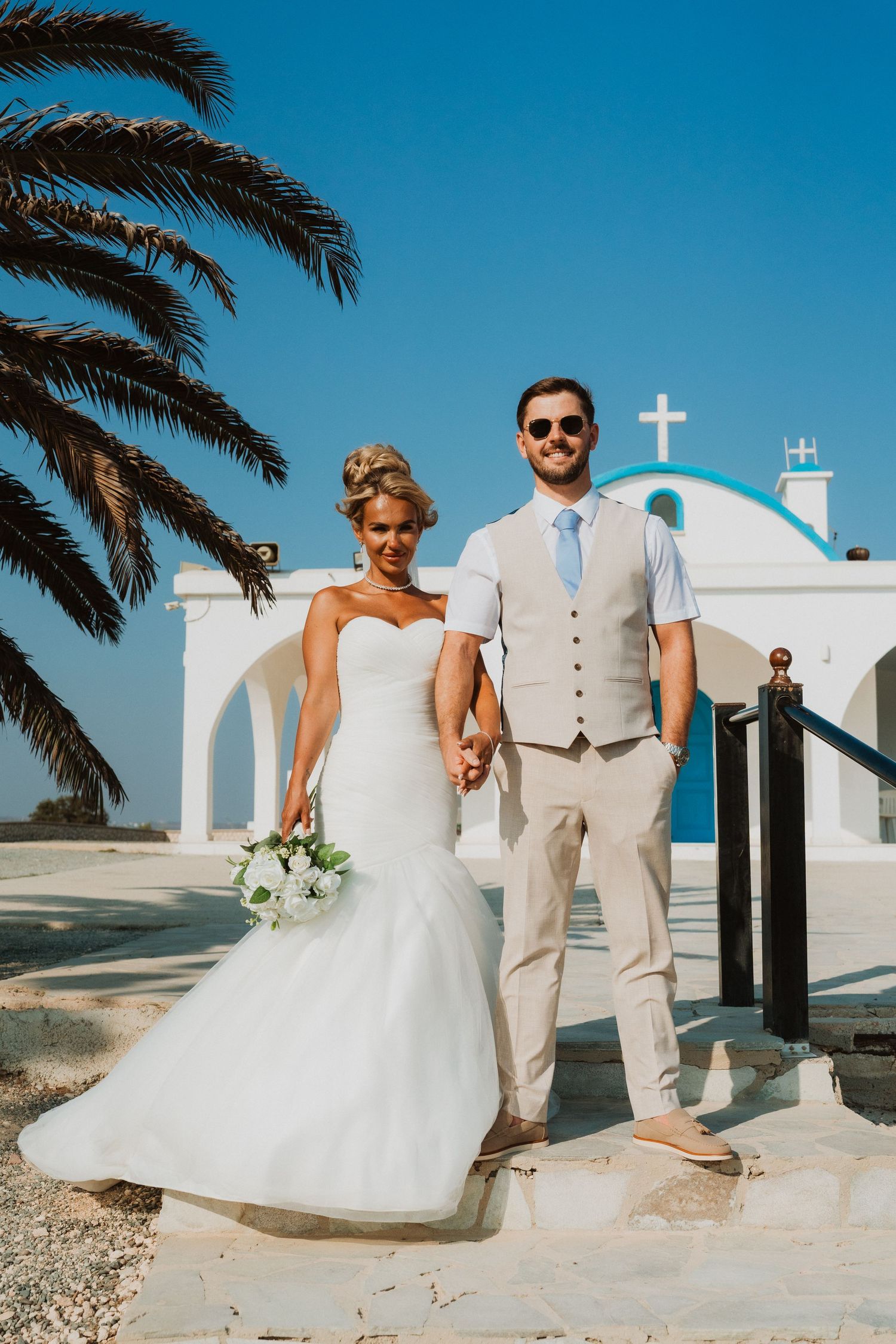 Bride and groom holding hands outside a white Greek chapel with blue dome and palm tree on sunny day.