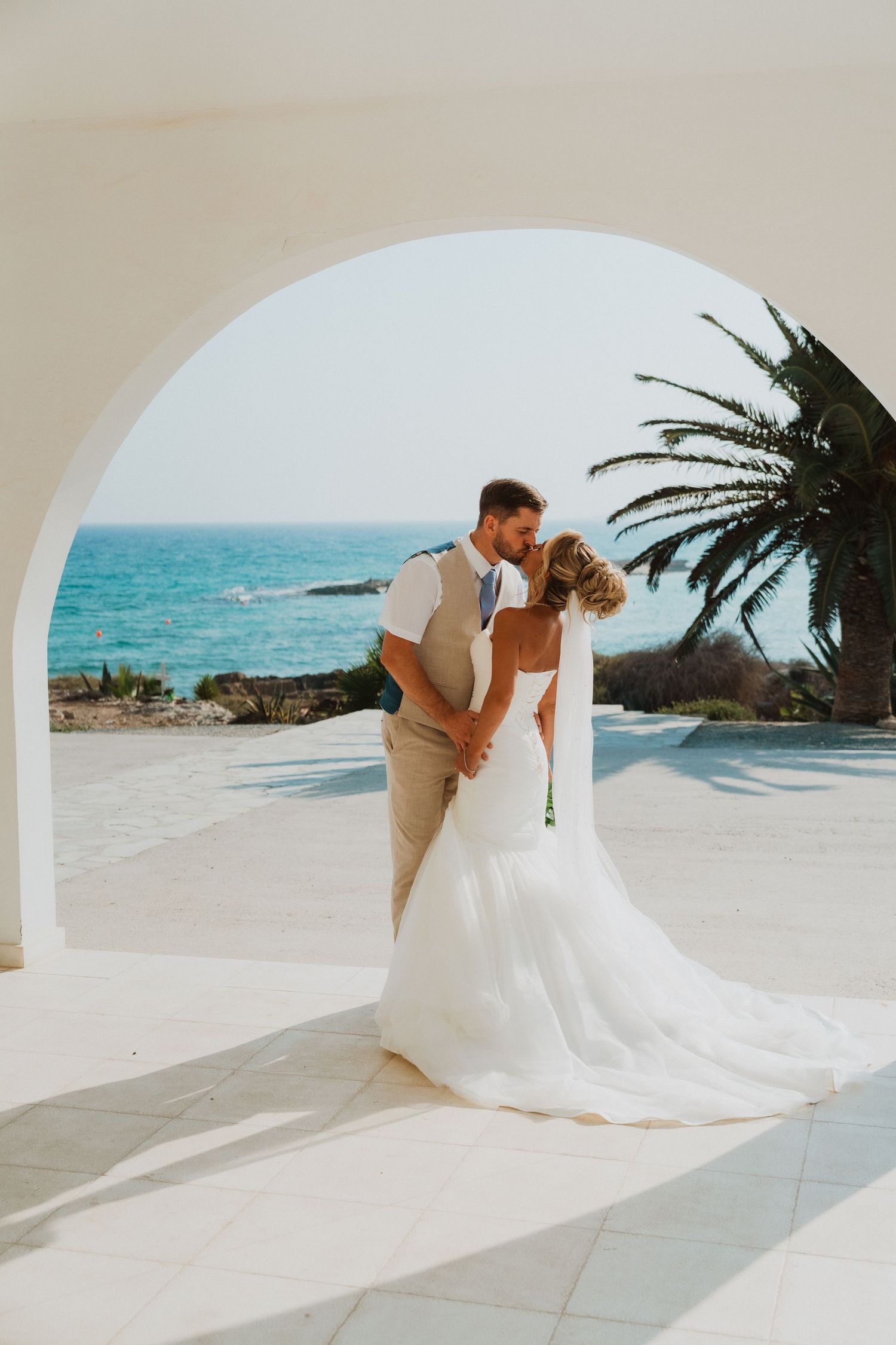 Bride and groom share a kiss under a white arch with turquoise ocean and palm tree backdrop at a beach wedding.