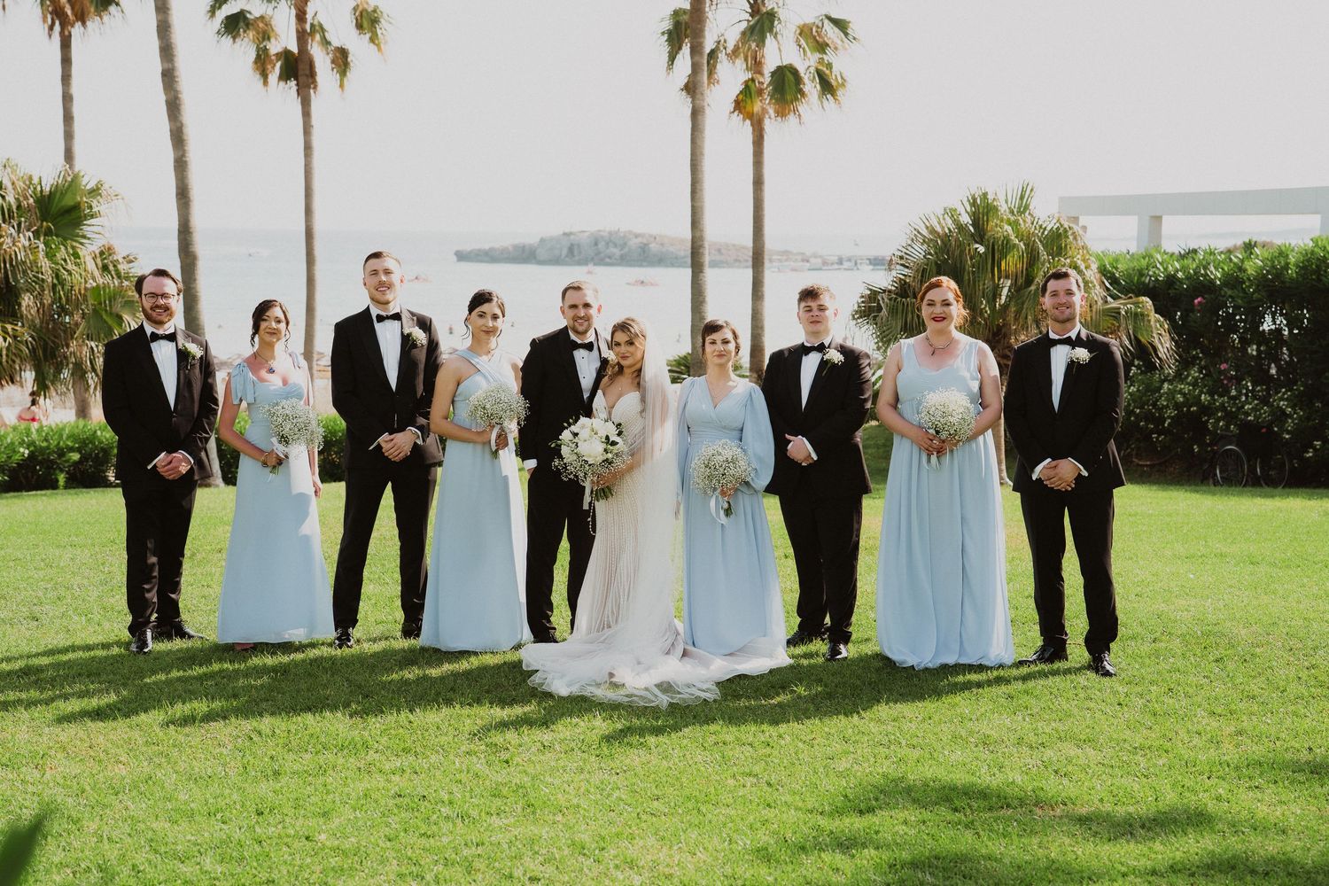 Wedding party poses on green lawn with bridesmaids in light blue gowns and groomsmen in black tuxedos by water.