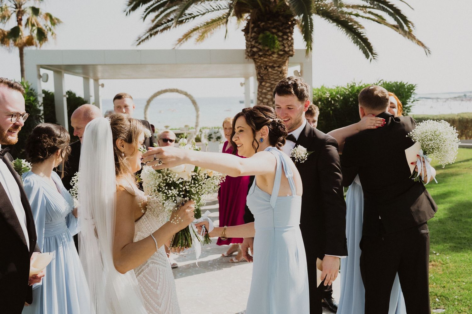 Bride and groom greet wedding guests with handshakes and hugs after outdoor ceremony near palm trees.