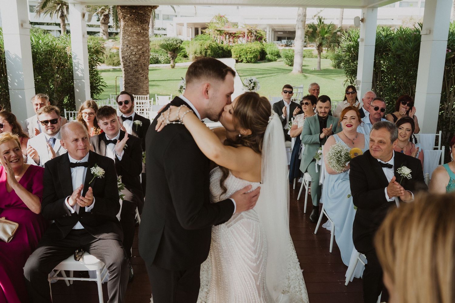 Bride and groom share first kiss at outdoor wedding ceremony as guests applaud in celebration.