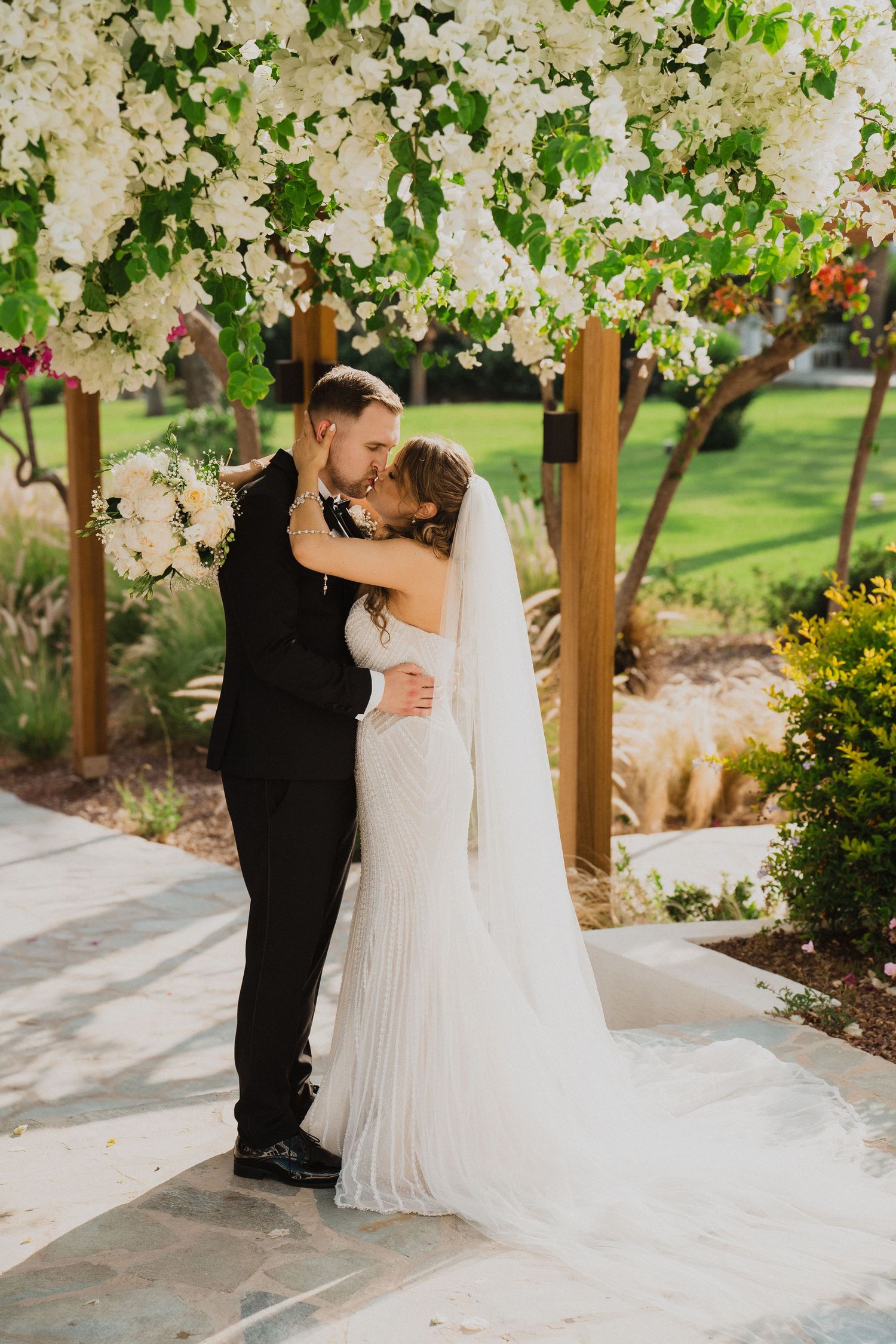 Bride and groom share a romantic kiss under a white floral pergola at an outdoor garden wedding.
