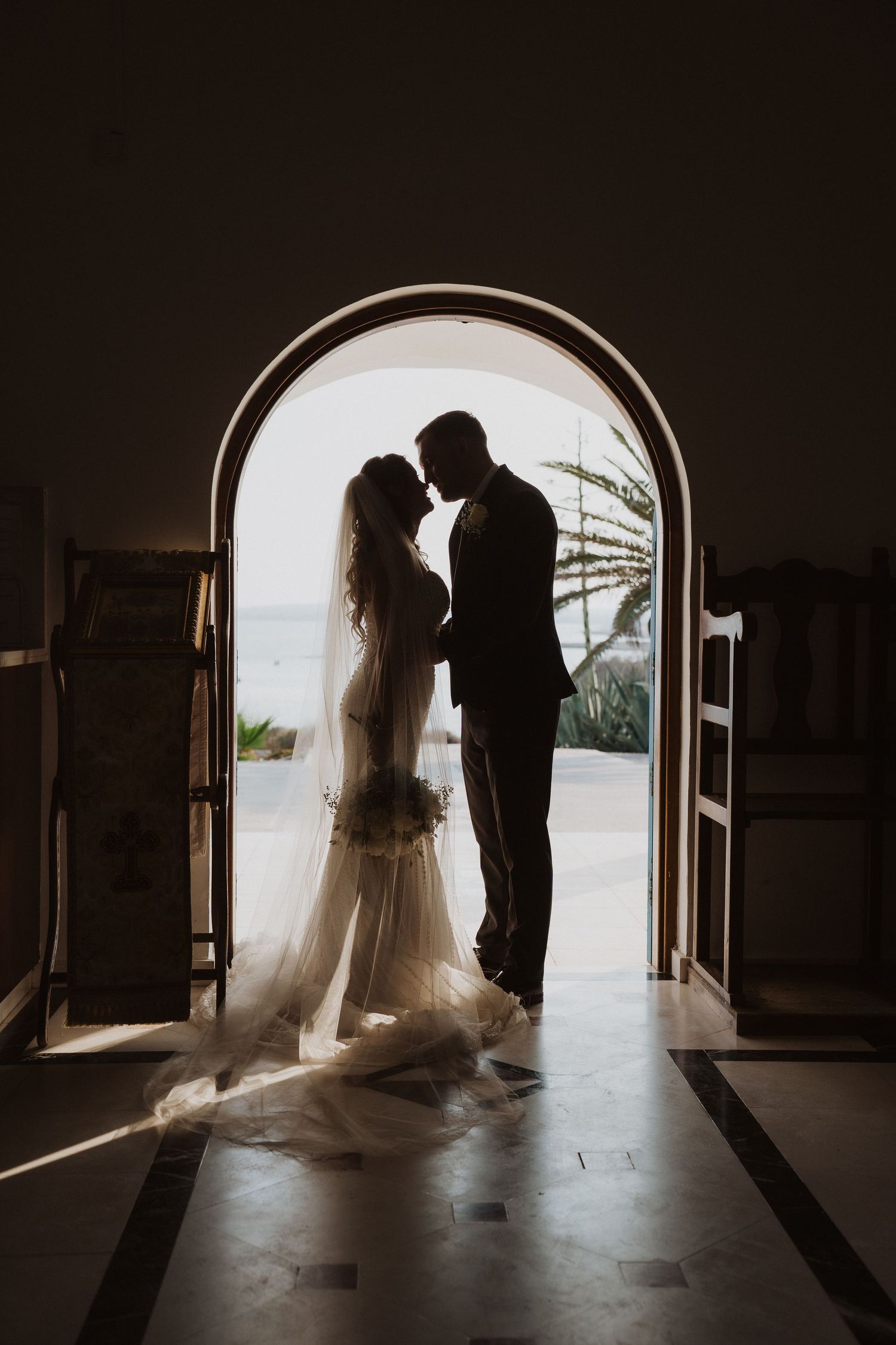 Dramatic silhouette of bride and groom embracing in an arched church doorway with ocean view.