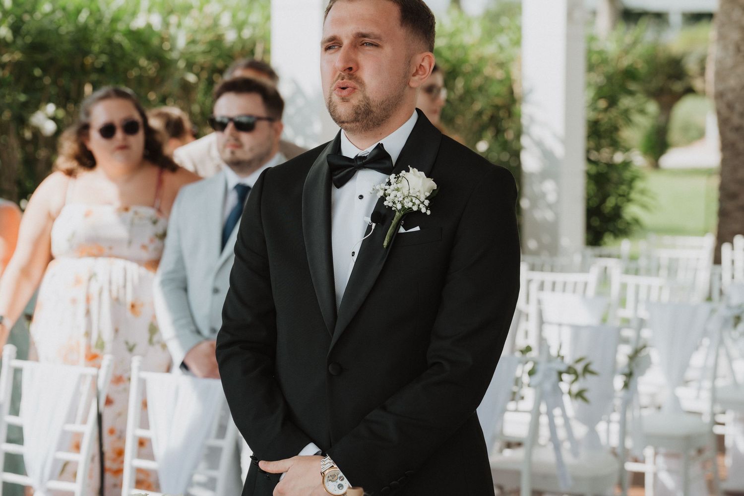 Emotional groom in black tuxedo waiting at the altar during an outdoor wedding ceremony.