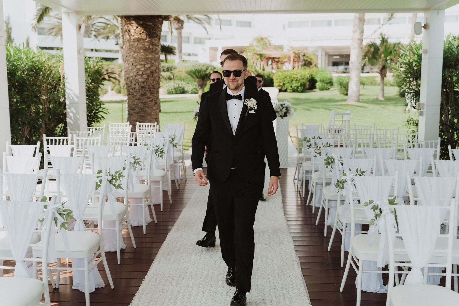 Dapper groom in black tuxedo walking down the aisle at an outdoor wedding ceremony.