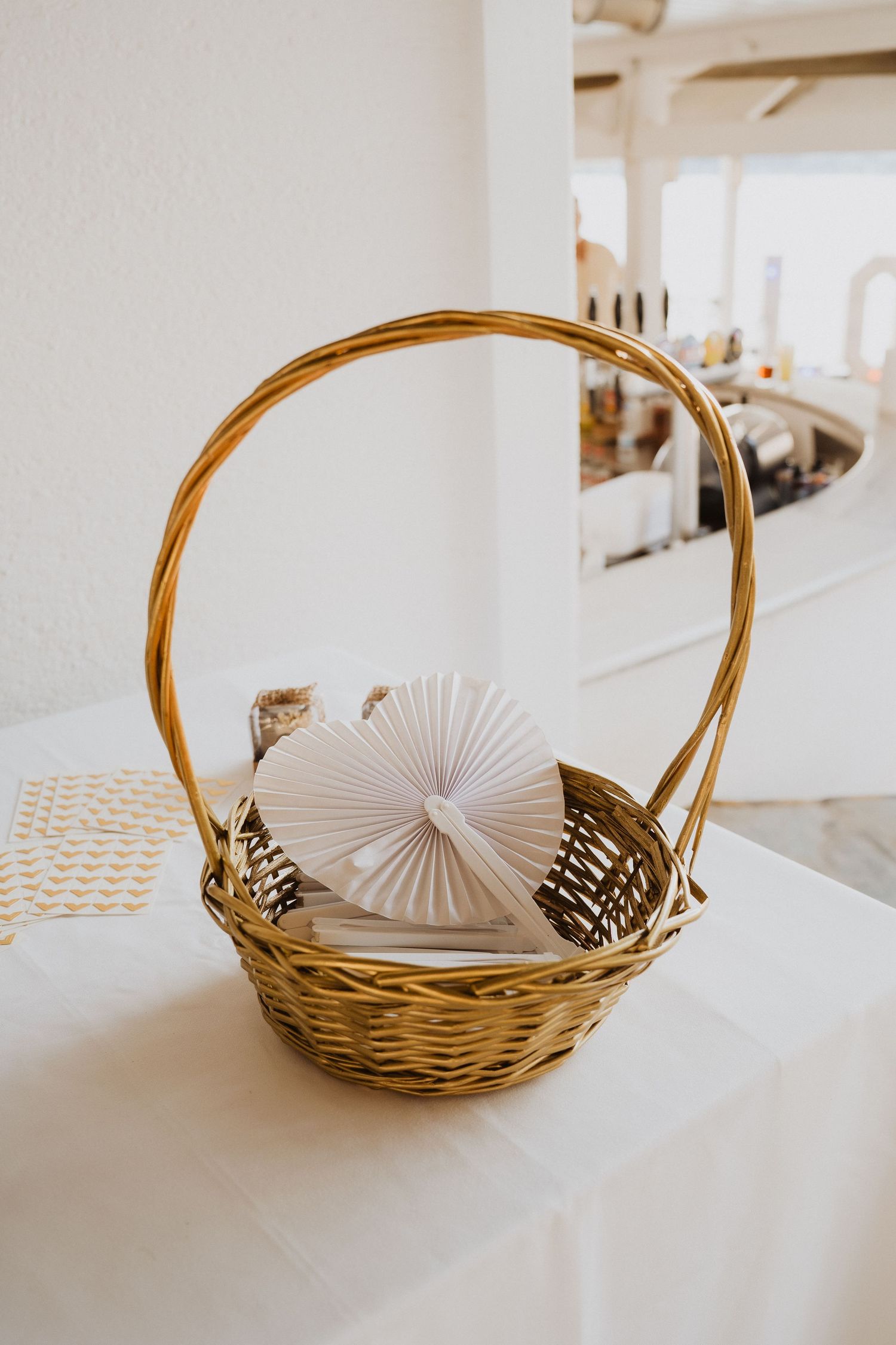 Wicker basket with white paper fan decoration sits on white tablecloth at elegant wedding reception venue.