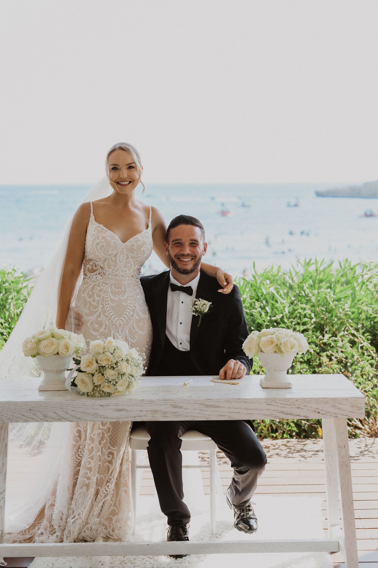 Newlyweds sharing joyful moments on white bench at seaside wedding venue.