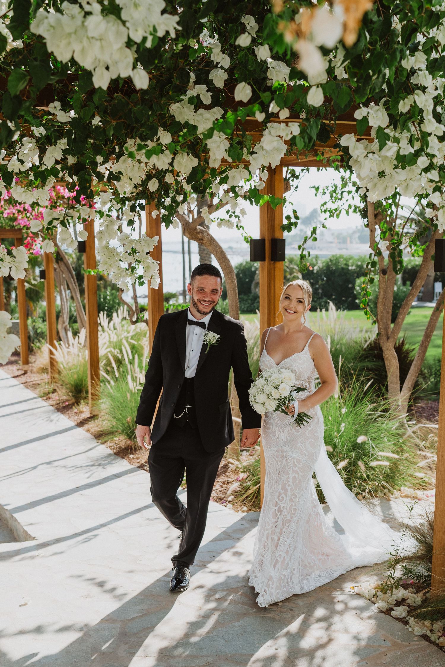 A couple poses under a beautiful white floral arbor in a garden setting on their wedding day.