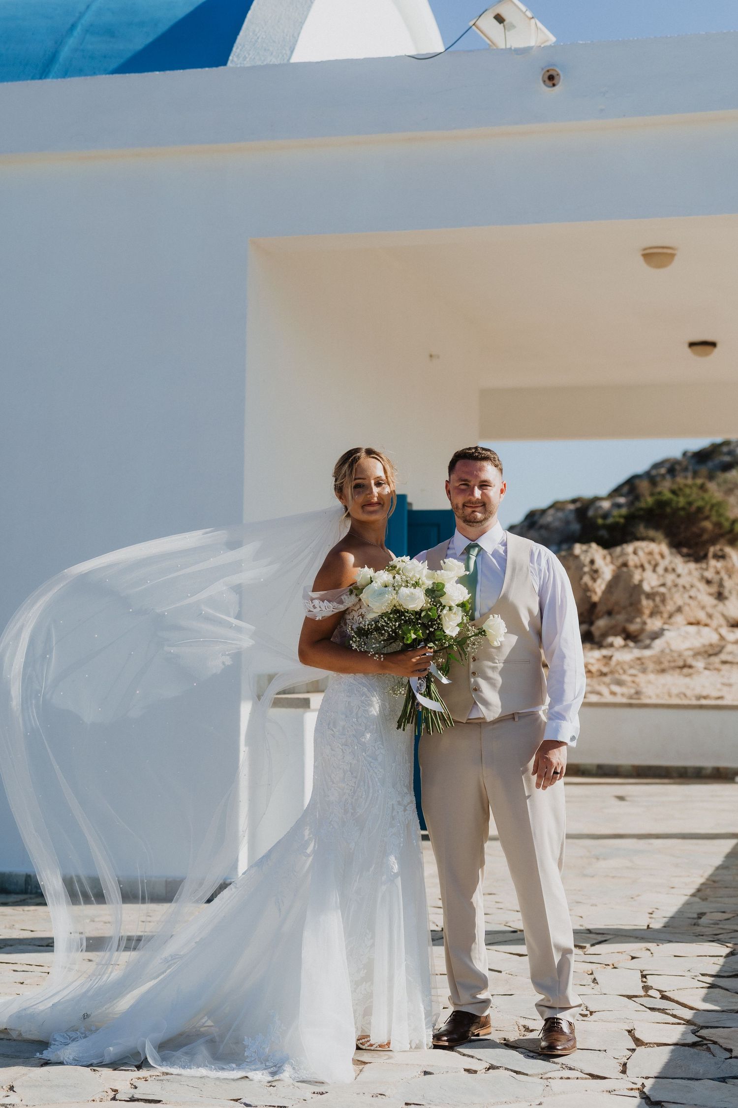 Newlywed couple in white attire walking on beach as bride's veil flows dramatically in the ocean breeze.