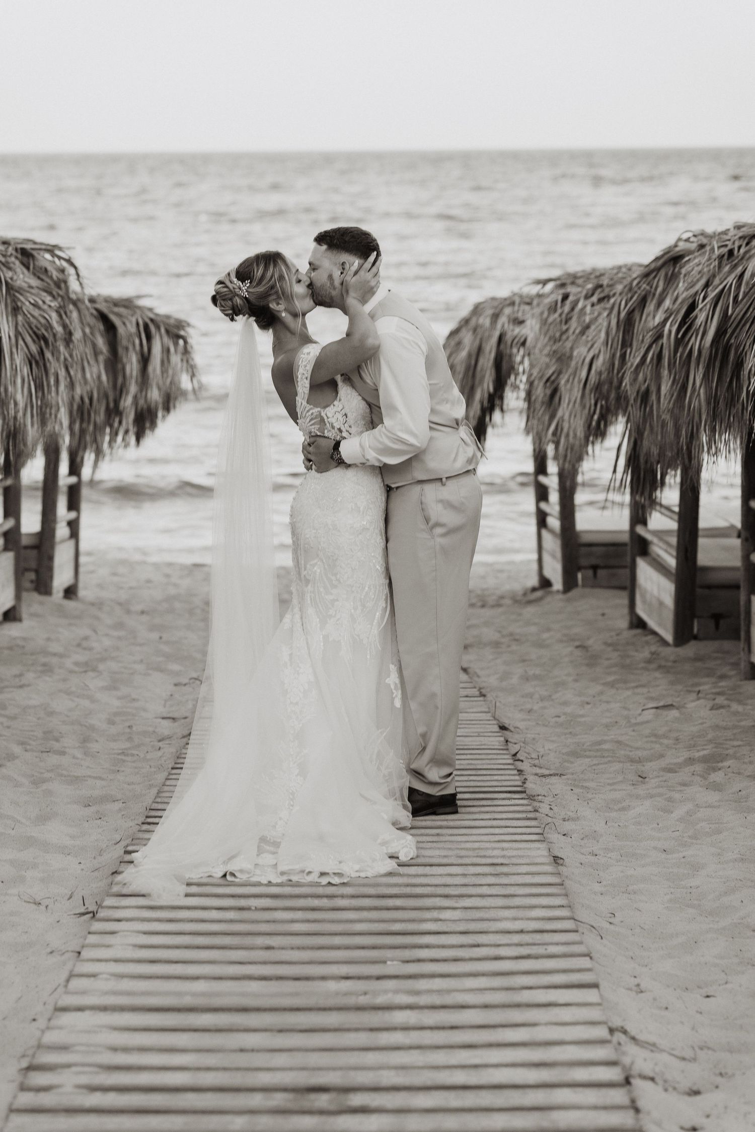 A romantic beach wedding scene with a couple embracing on a wooden boardwalk near thatched umbrellas.