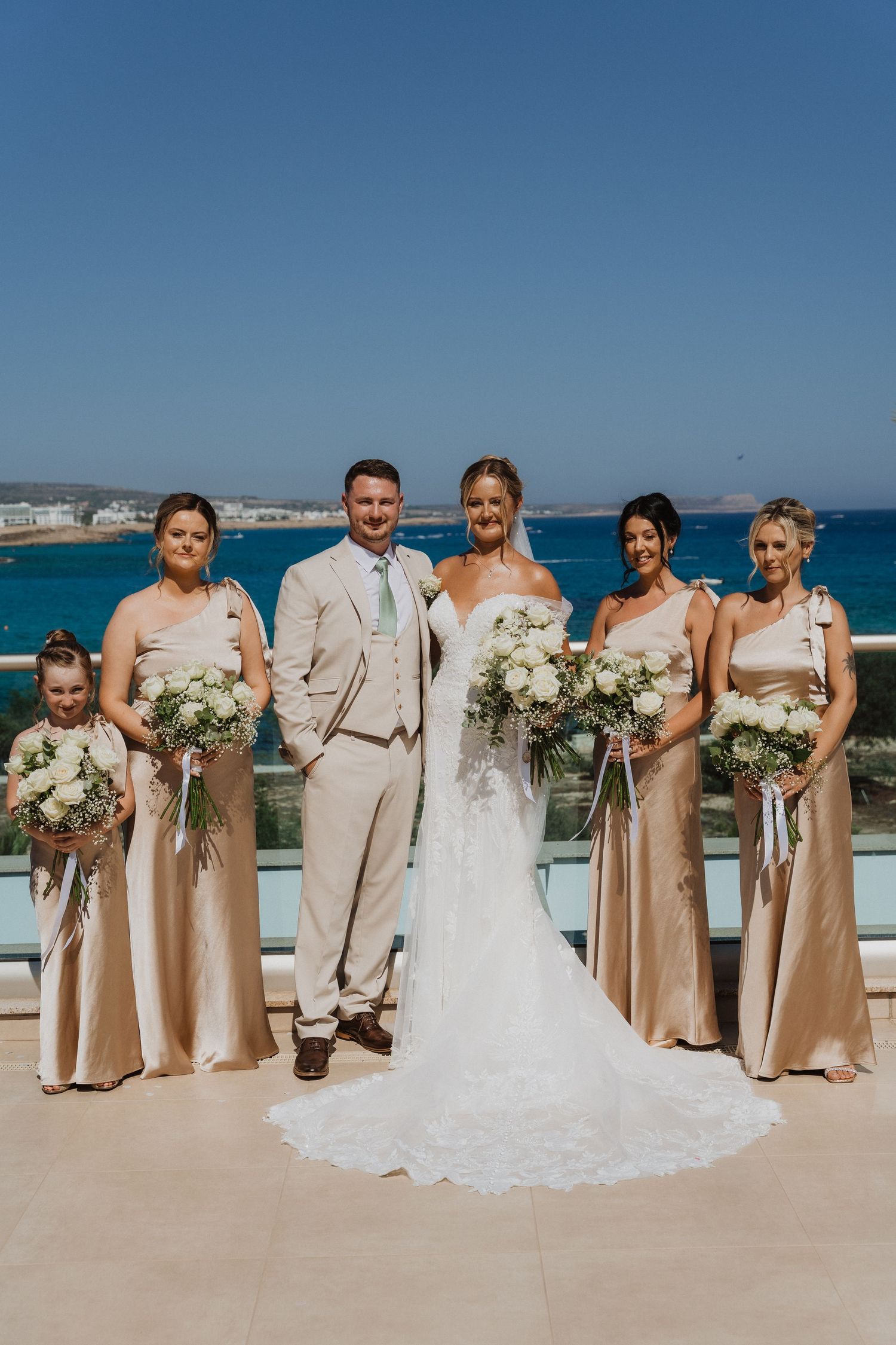 Wedding party in beige and white attire poses together on a sunny beach with ocean views in the background.