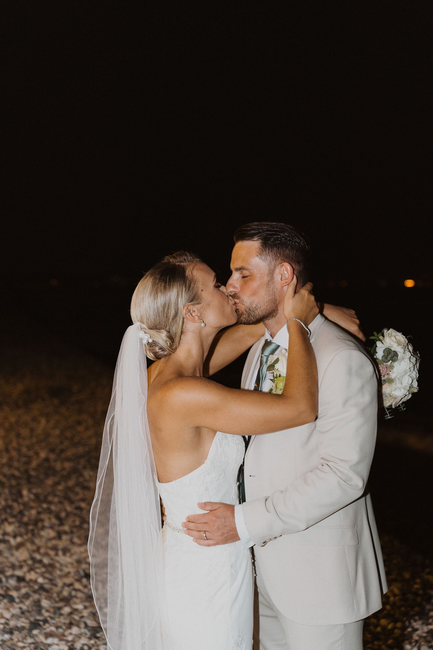 Newlyweds share a passionate kiss during their nighttime beach wedding celebration.