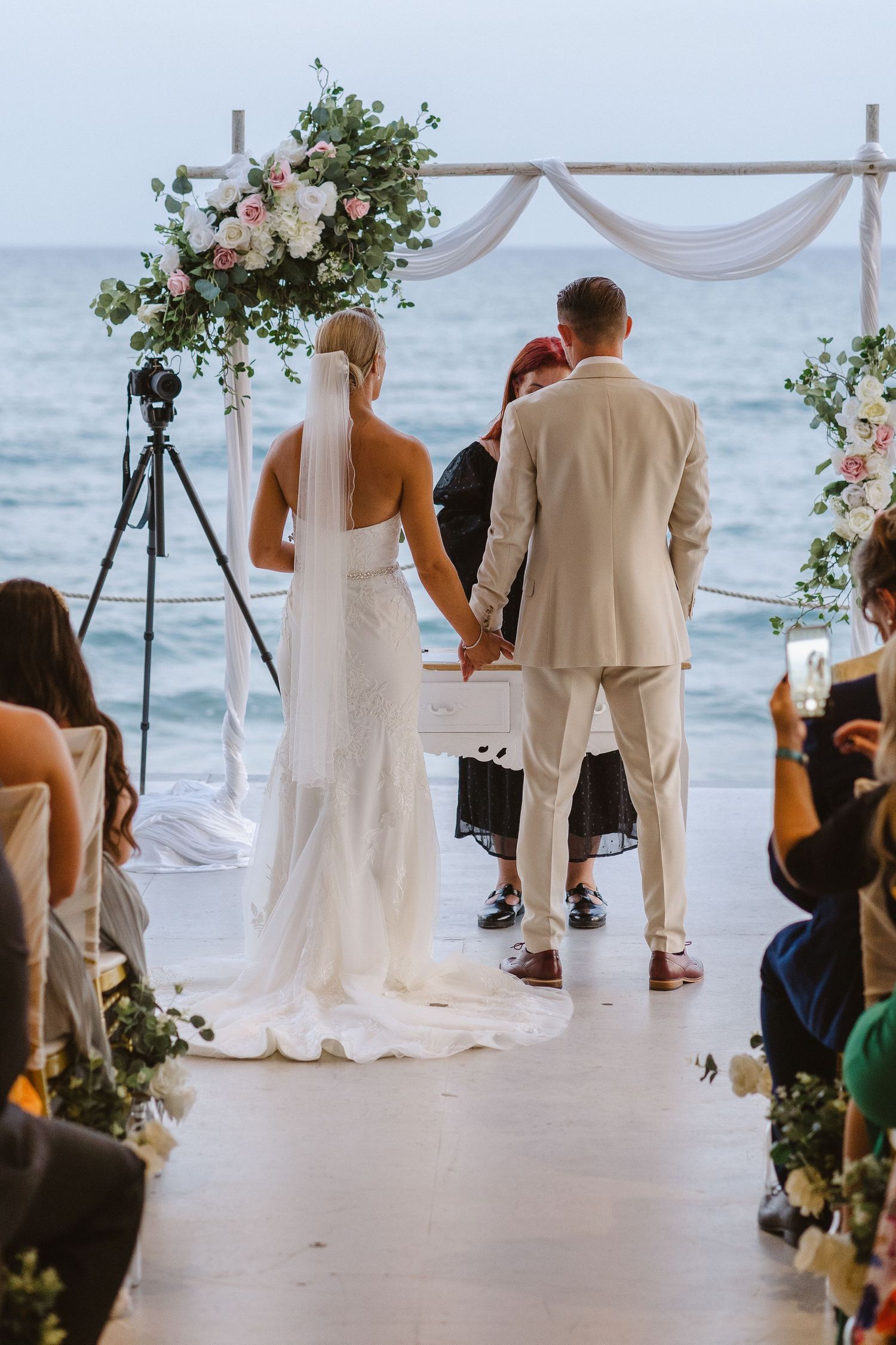 Oceanfront wedding ceremony with floral arch and guests seated under covered pavilion.