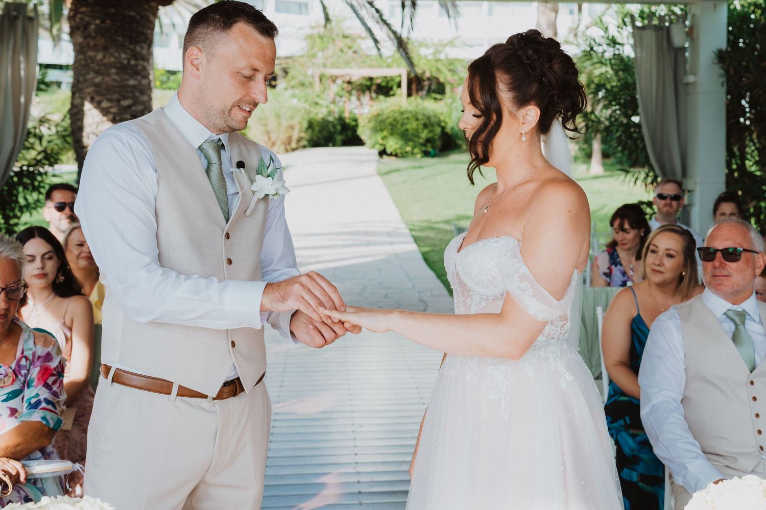 A couple exchanges rings during an outdoor wedding ceremony while guests watch from their seats.
