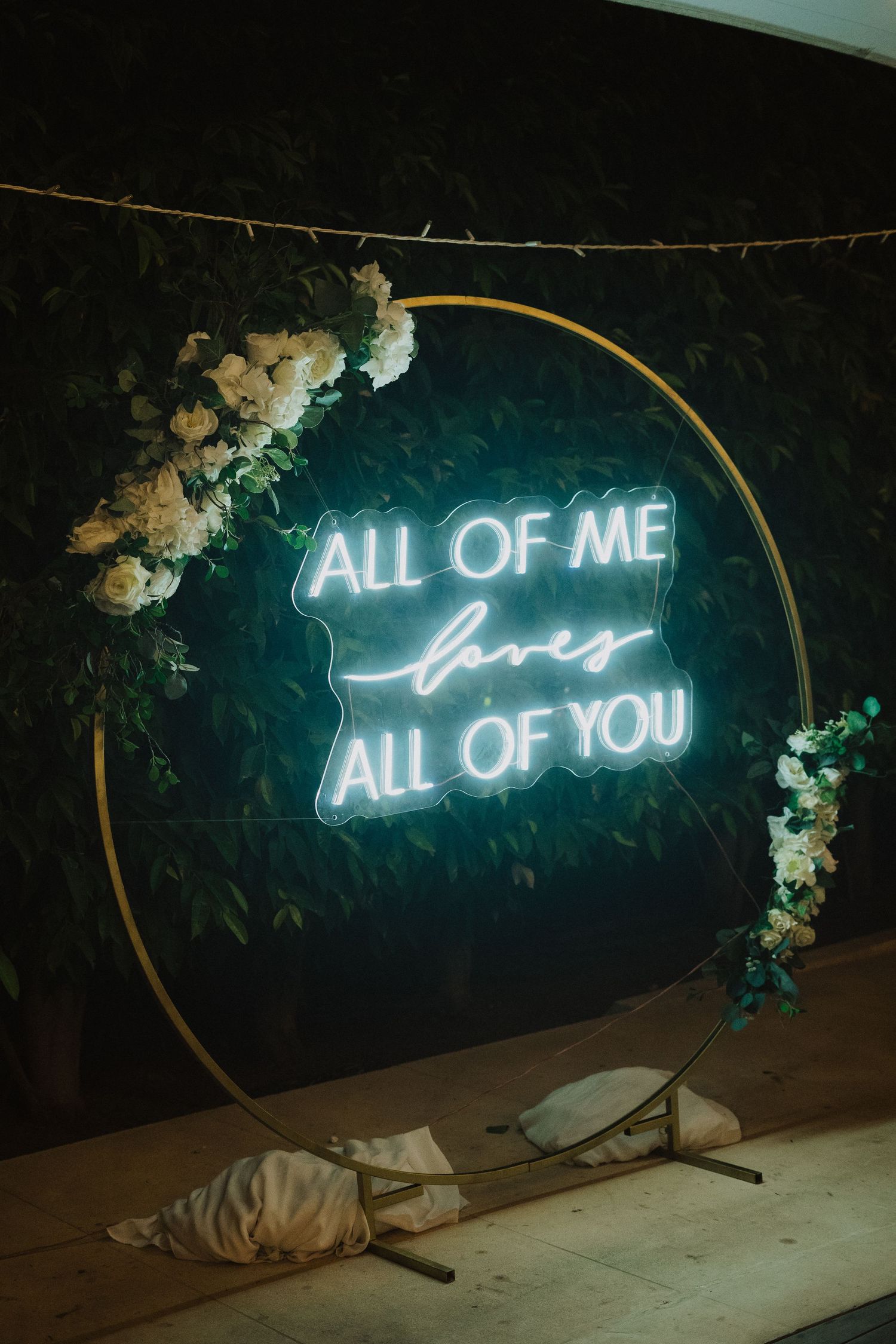 A neon sign reading 'All of me loves all of you' mounted on a dark wall with floral decorations.