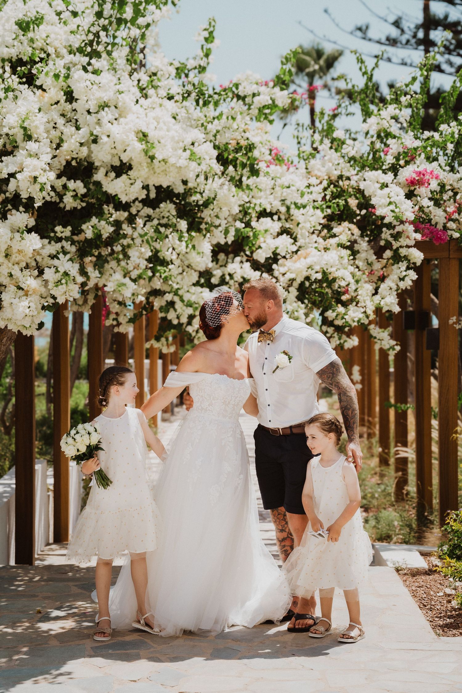 Newlyweds share a moment under blooming white flowers while two girls in white dresses look on.