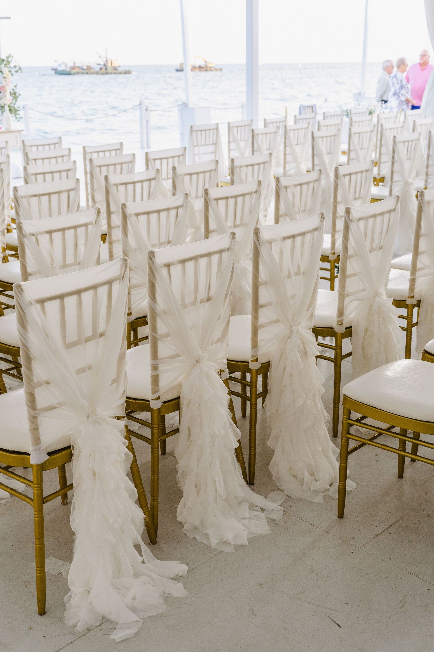 White chiavari chairs with flowing fabric decorations arranged for a waterfront ceremony.