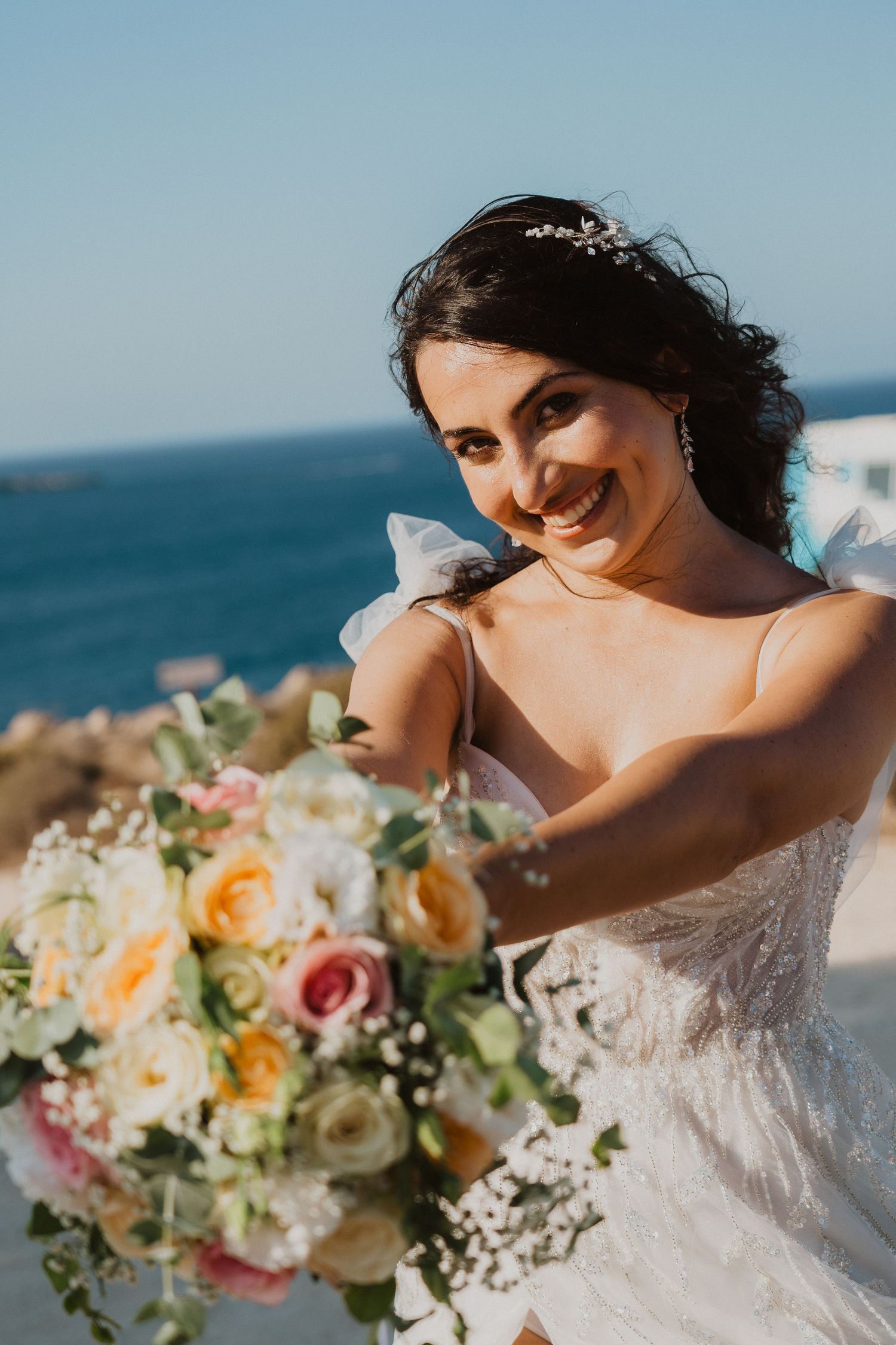 Beautiful bride in white lace dress holds colorful bouquet of roses and mixed flowers against ocean backdrop.