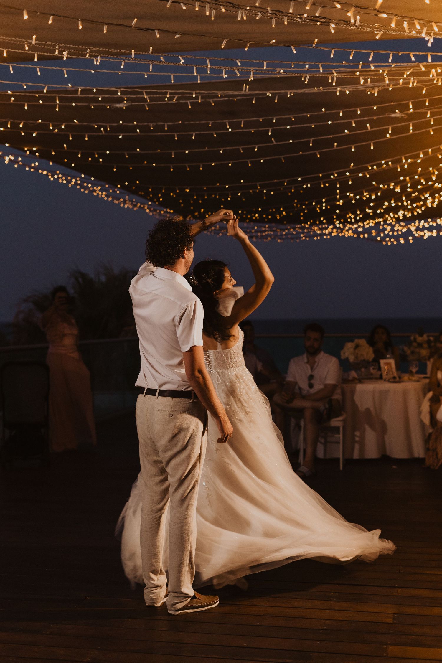 Elegant couple twirls on wooden dance floor beneath a magical ceiling of fairy lights at dusk.