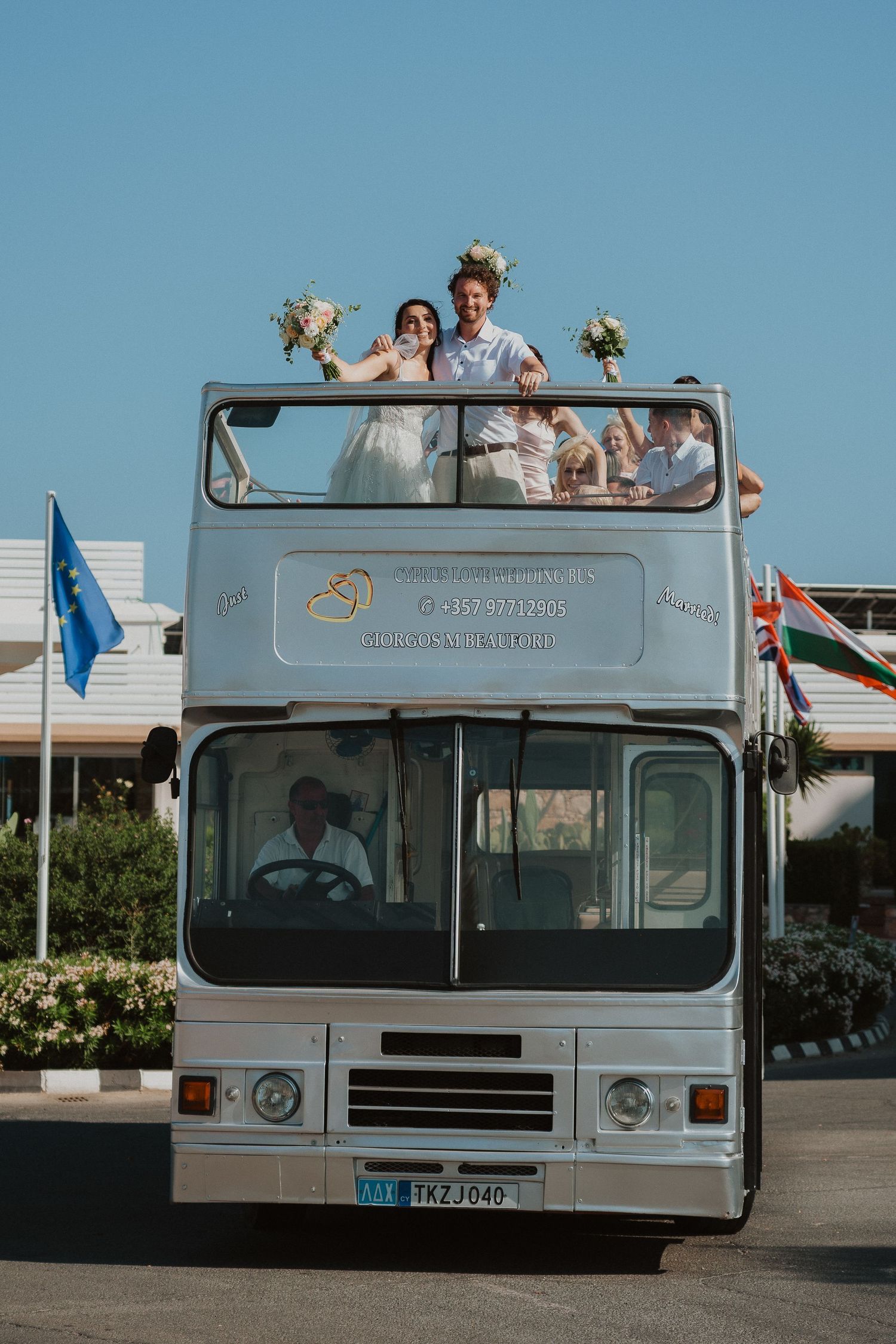 Newlyweds celebrating atop vintage double-decker bus against blue sky.