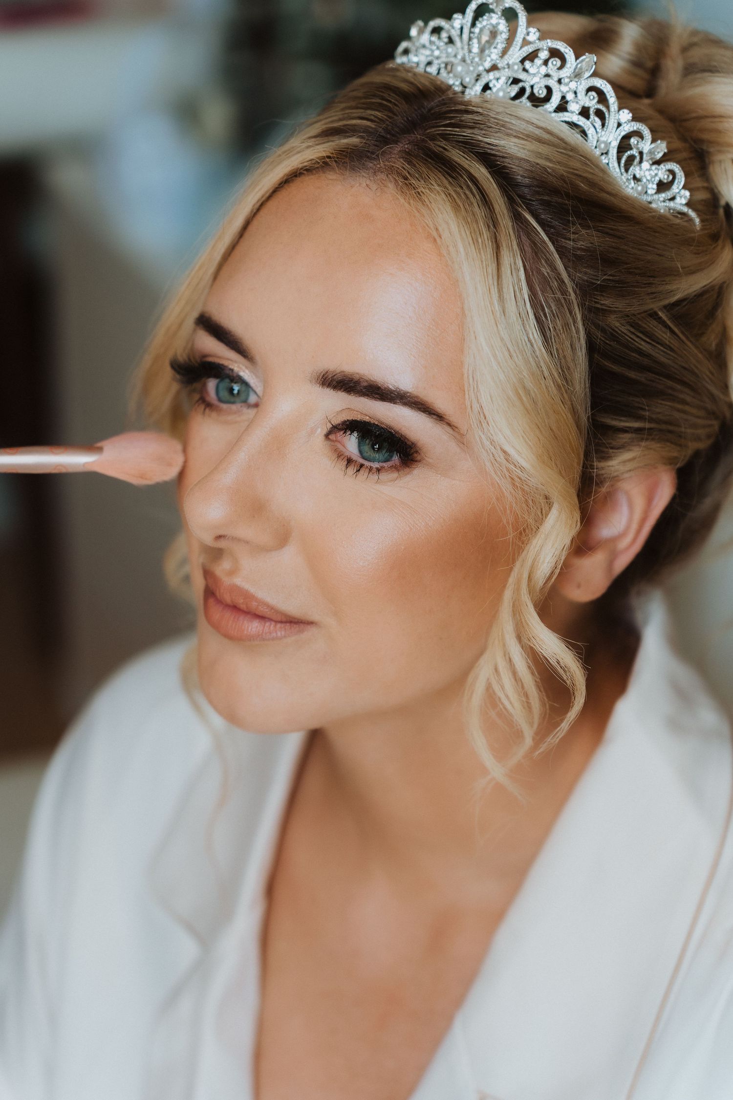 Elegant bridal makeup application with delicate crystal tiara and soft blonde updo in natural lighting.