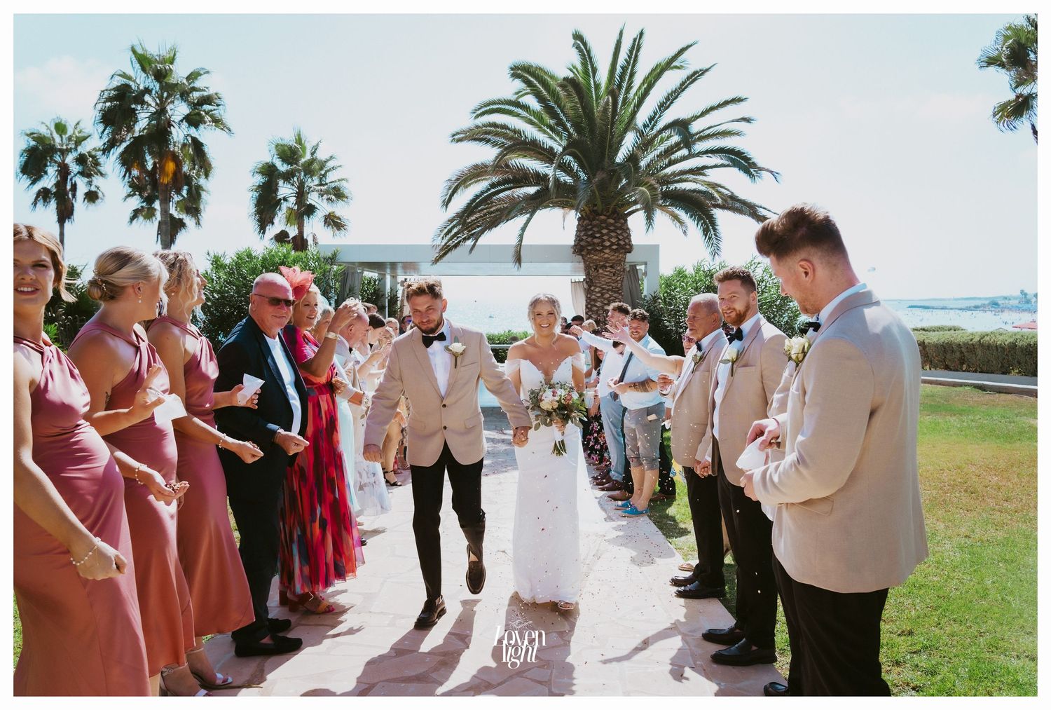 Wedding guests form an aisle under palm trees while celebrating the couple's exit.