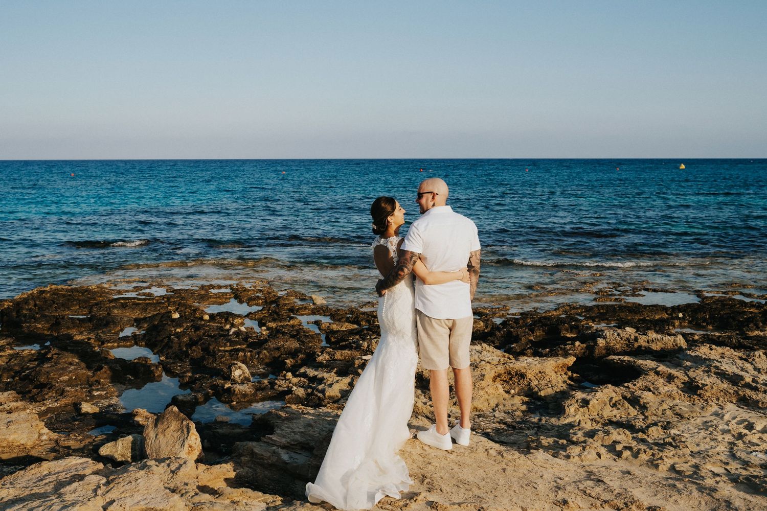 A wedding couple stands on rocky coastline with deep blue Mediterranean waters in the background.