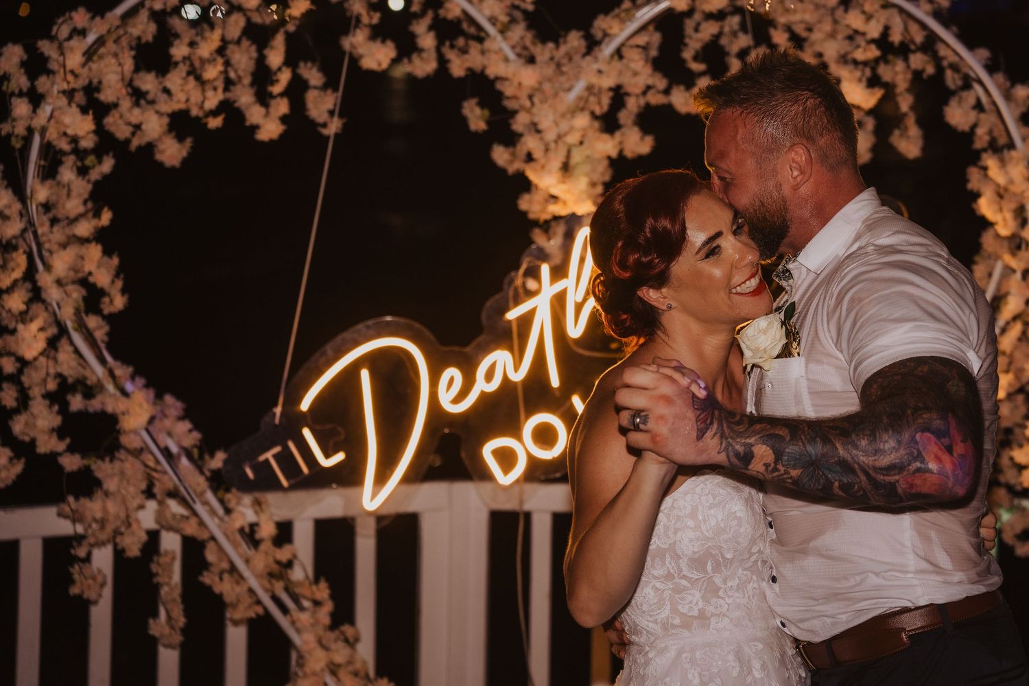 Couple embraces during first dance under illuminated neon sign at wedding reception.