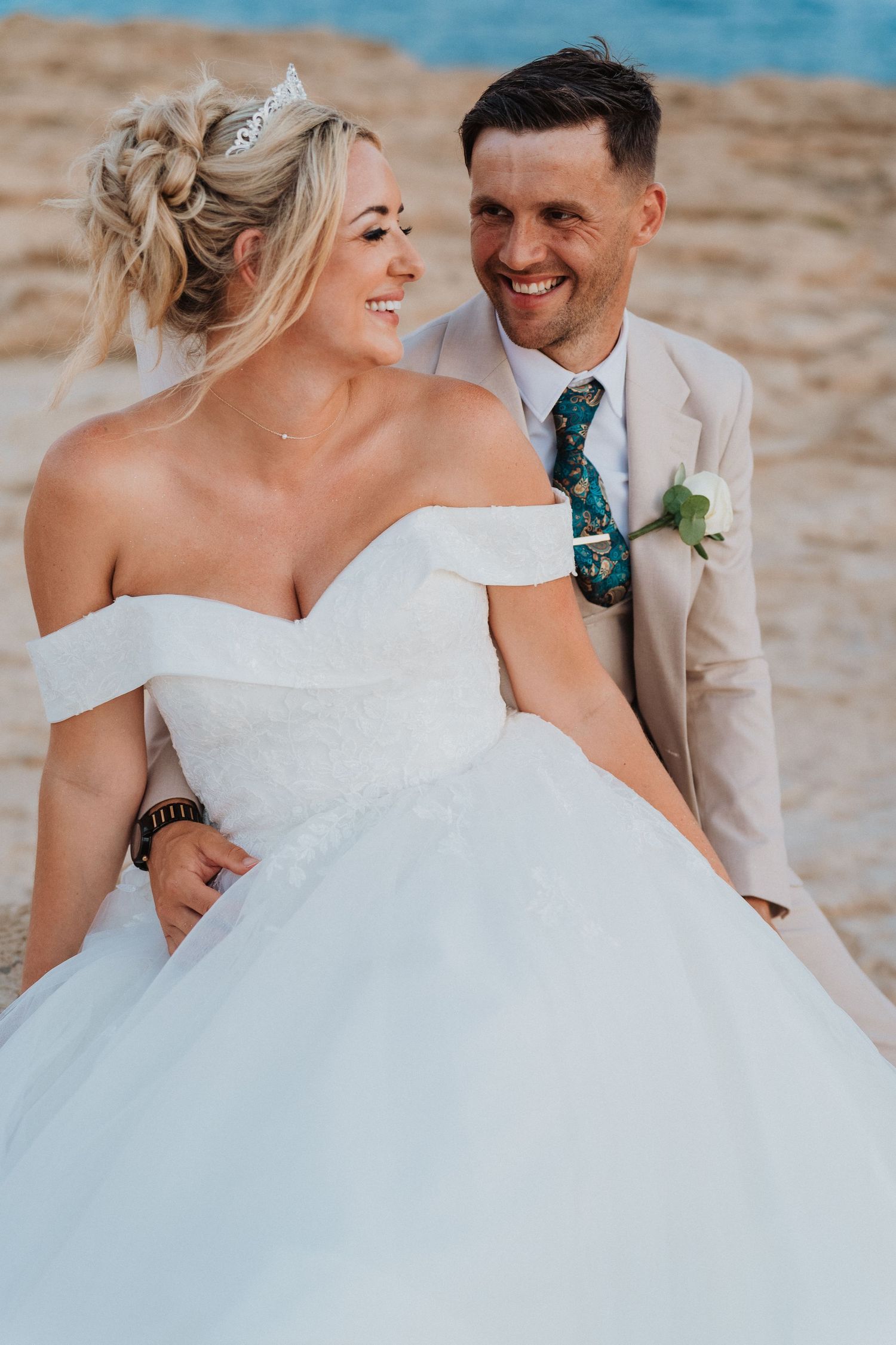 A joyous wedding couple share a candid moment on the beach, with the bride wearing an elegant off-shoulder white gown.