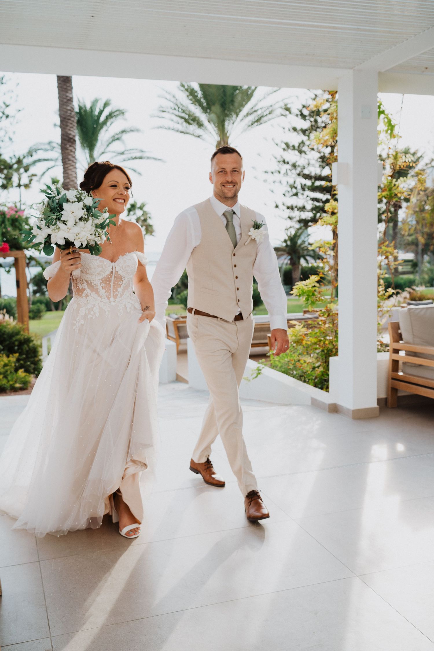 Happy newlyweds walking together on a covered porch, bride in white gown and groom in light-colored suit and vest.