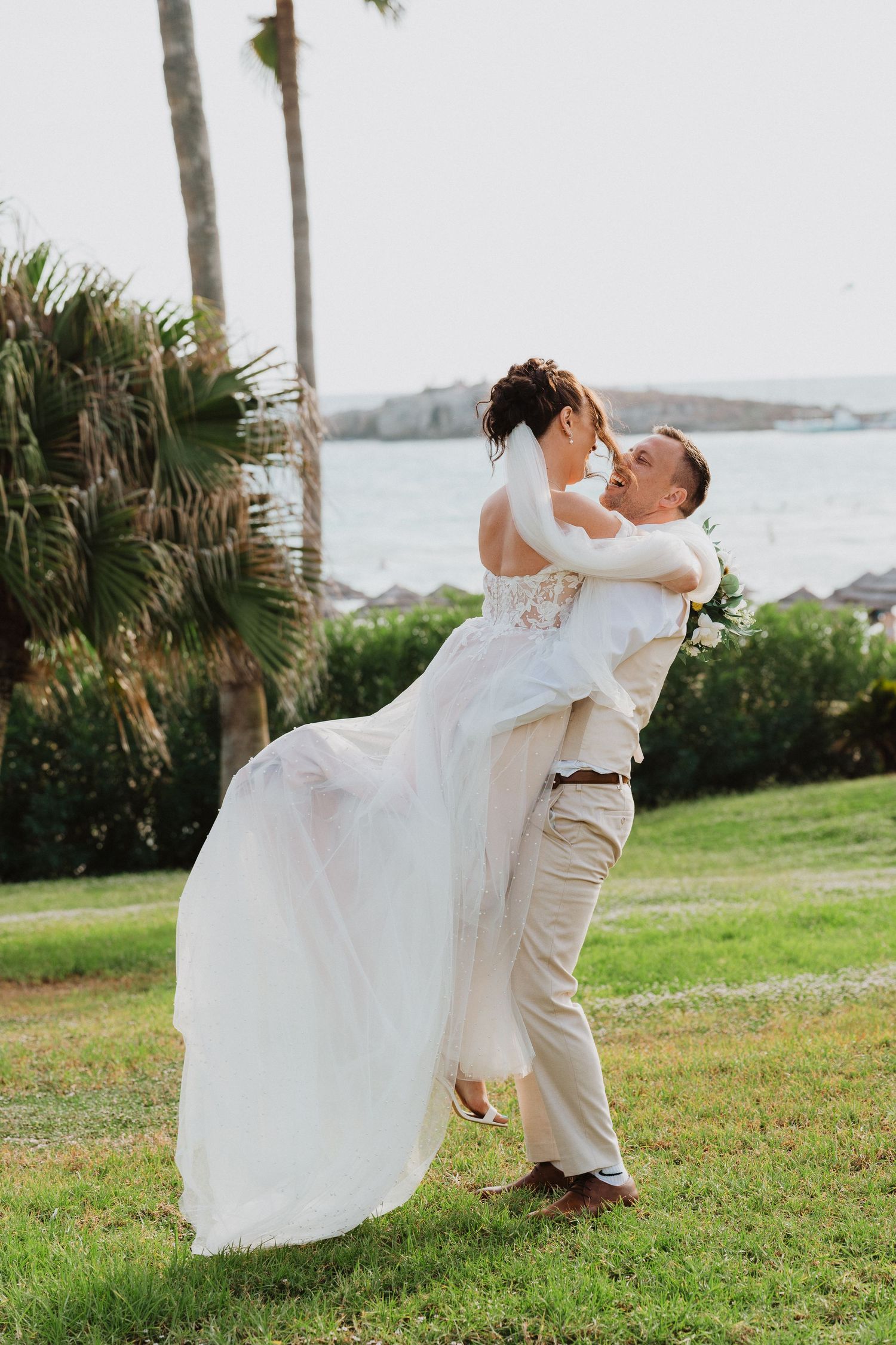A couple embraces during their outdoor tropical beach wedding, with palm trees and ocean views in the background.
