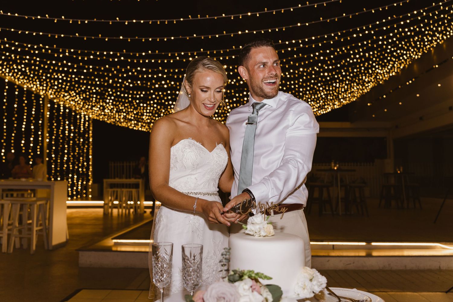 Newlyweds pose by their wedding cake under a romantic canopy of fairy lights.
