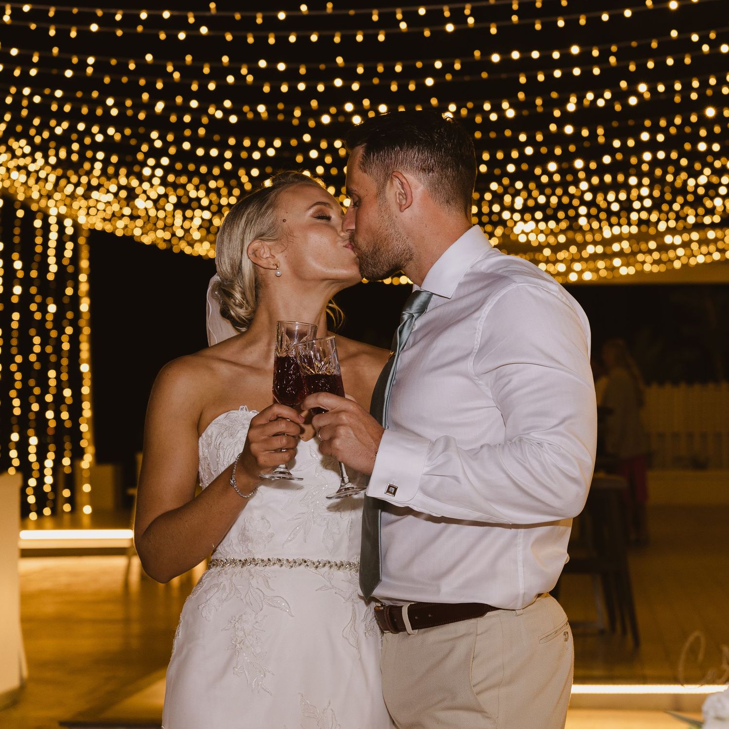 Newlyweds share a passionate kiss while holding champagne under twinkling fairy lights.