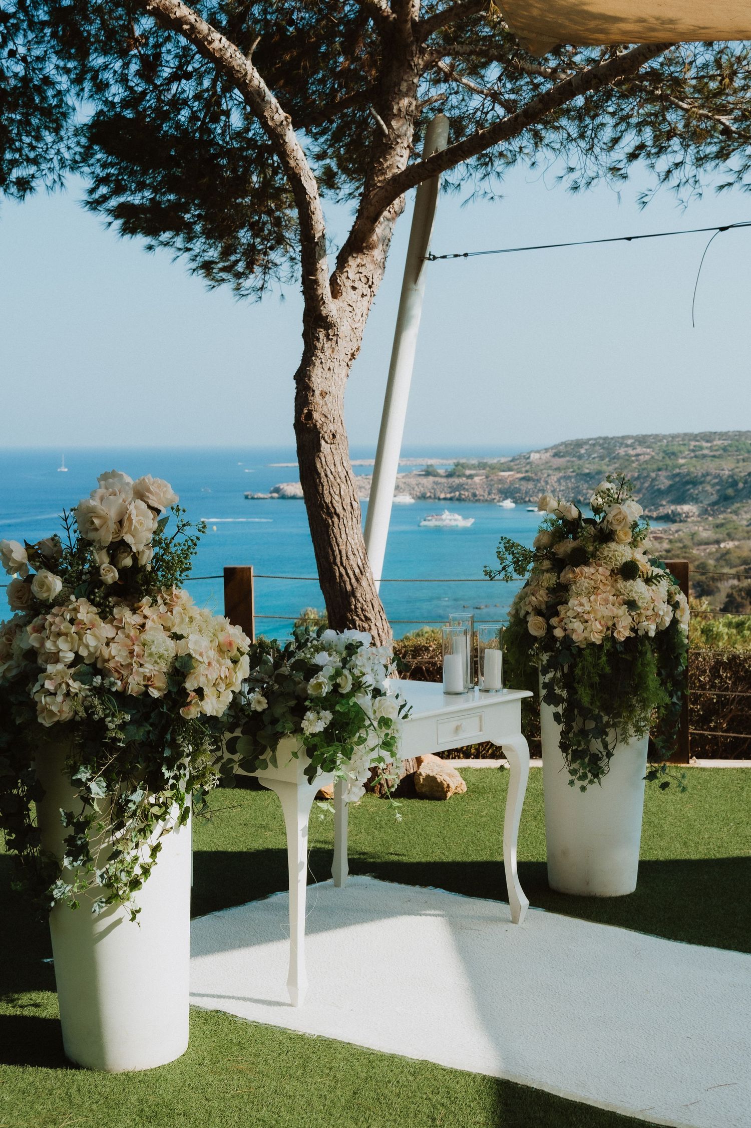 An outdoor wedding setup overlooking a Mediterranean coastline with white floral arrangements and a pine tree.